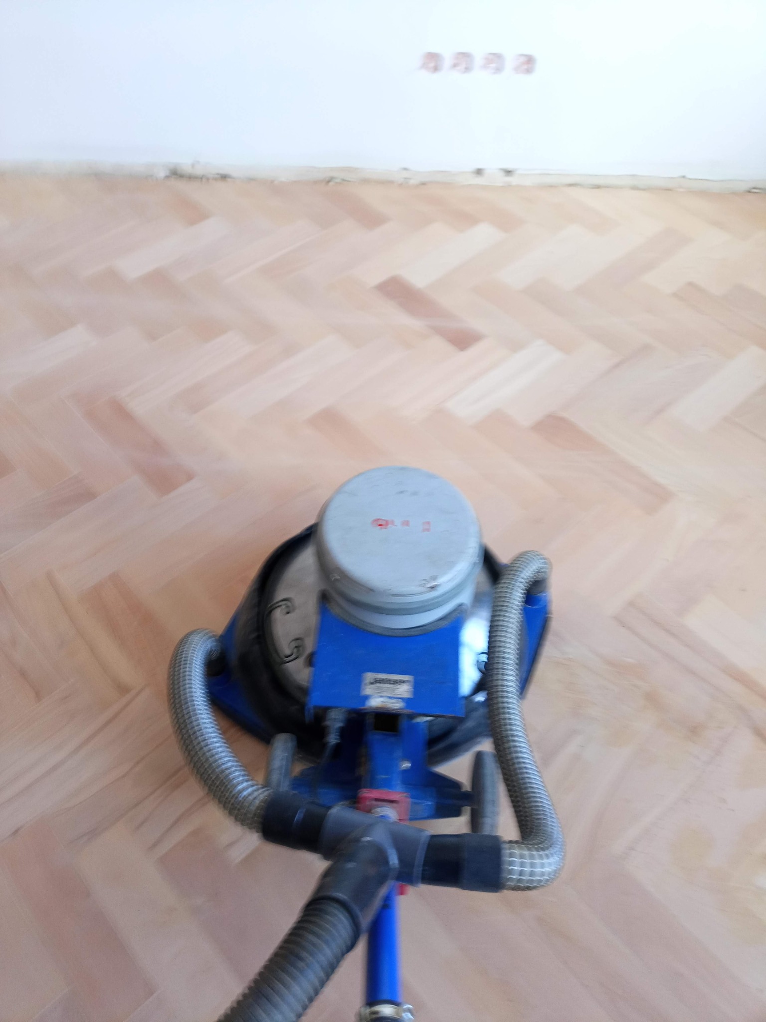 Widok z góry na maszynę do obróbki parkietu na tle jasnej, ułożonej w jodełkę podłogi parkietowej, z widocznymi gniazdkami elektrycznymi na ścianie w tle.