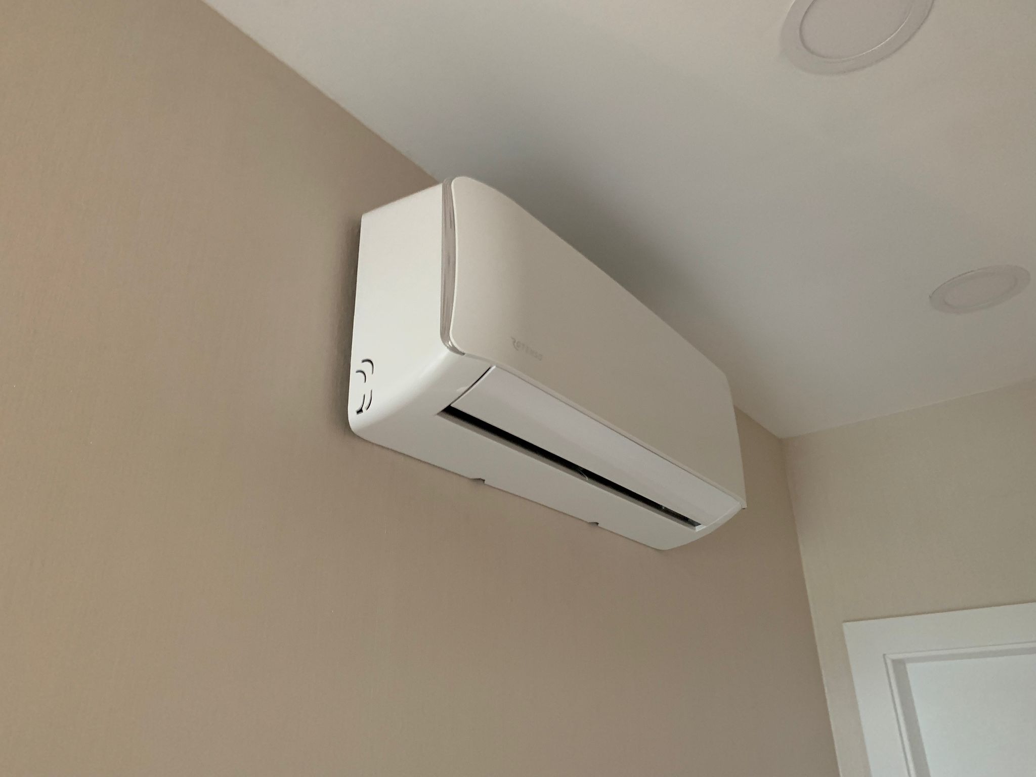 Biała klimatyzacja Rotenso zamontowana na beżowej ścianie w pomieszczeniu z białym sufitem i wbudowanymi lampami, widoczny fragment białych drzwi.