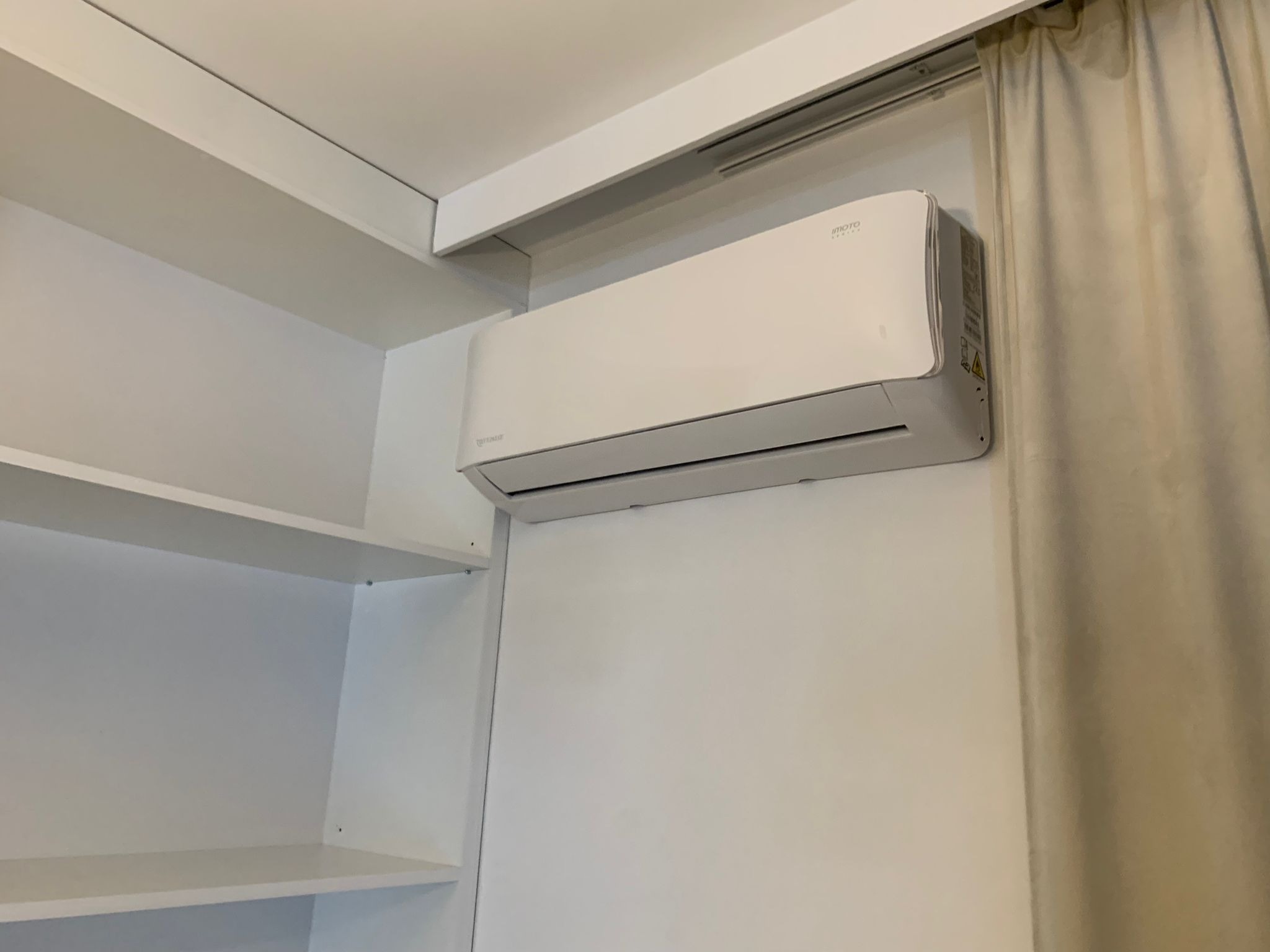 Biały klimatyzator ścienny marki Imoto zamontowany w pomieszczeniu z białymi meblami i zasłoną w tle. Widoczny z boku.