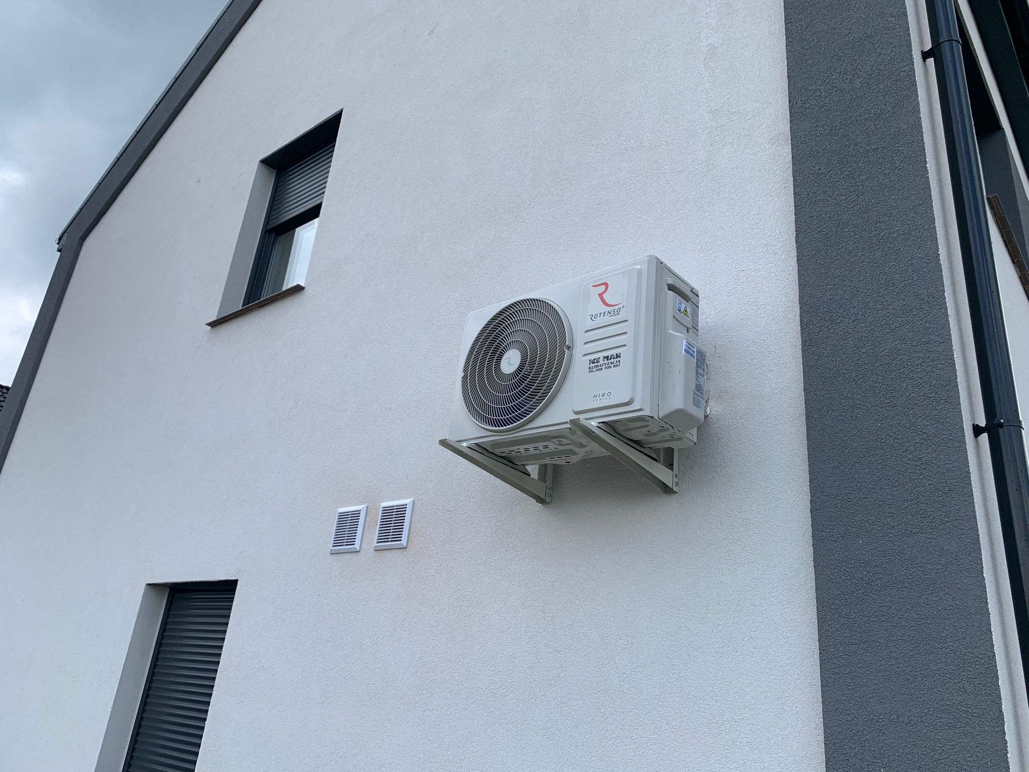 Zewnętrzna jednostka klimatyzacji Rotenso zamontowana na elewacji nowoczesnego domu z szarą stolarką okienną i dwoma kratkami wentylacyjnymi obok.