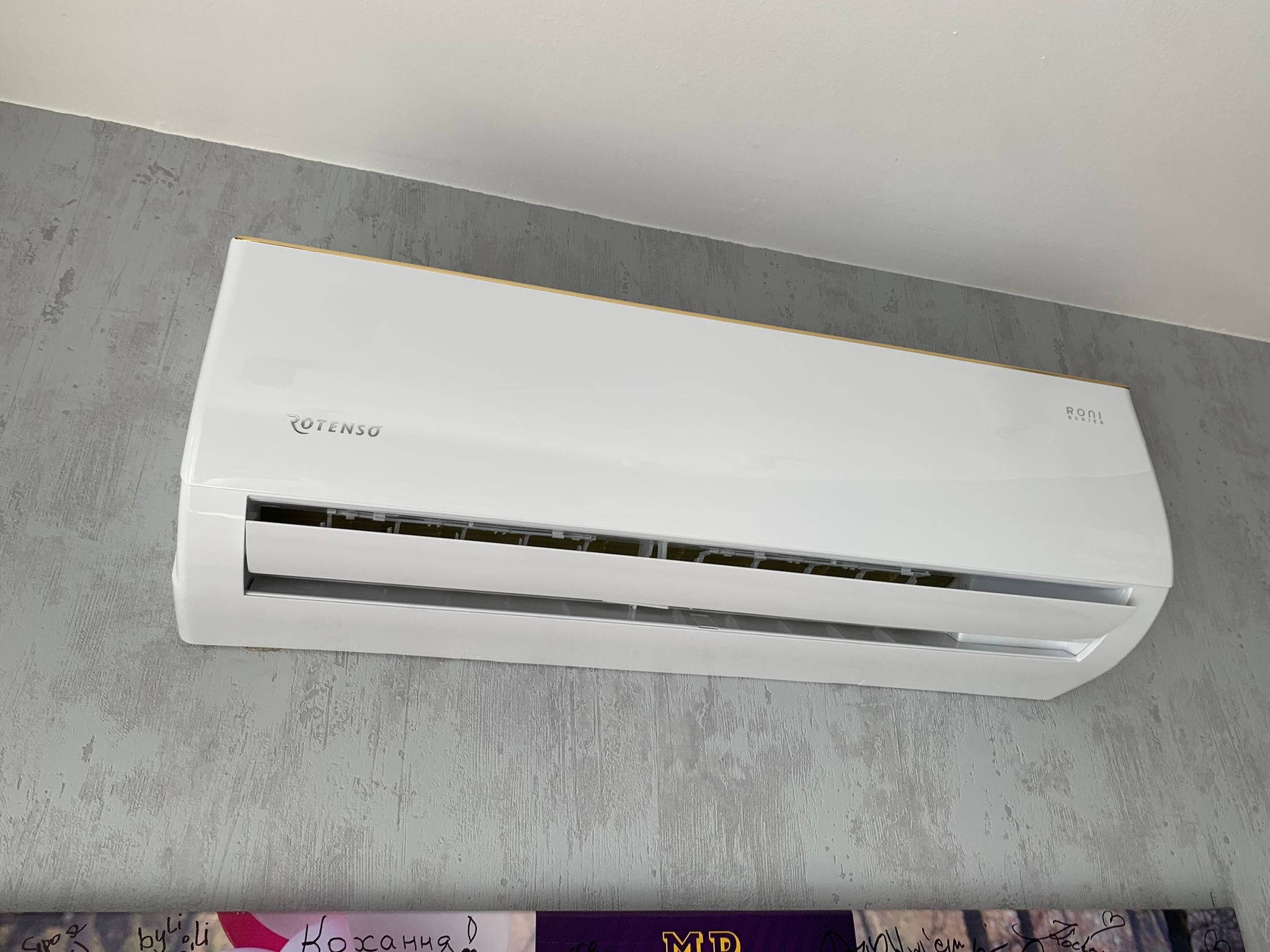 Biały klimatyzator Rotenso zamontowany na szarej ścianie z dekoracyjnymi napisami i rysunkami.