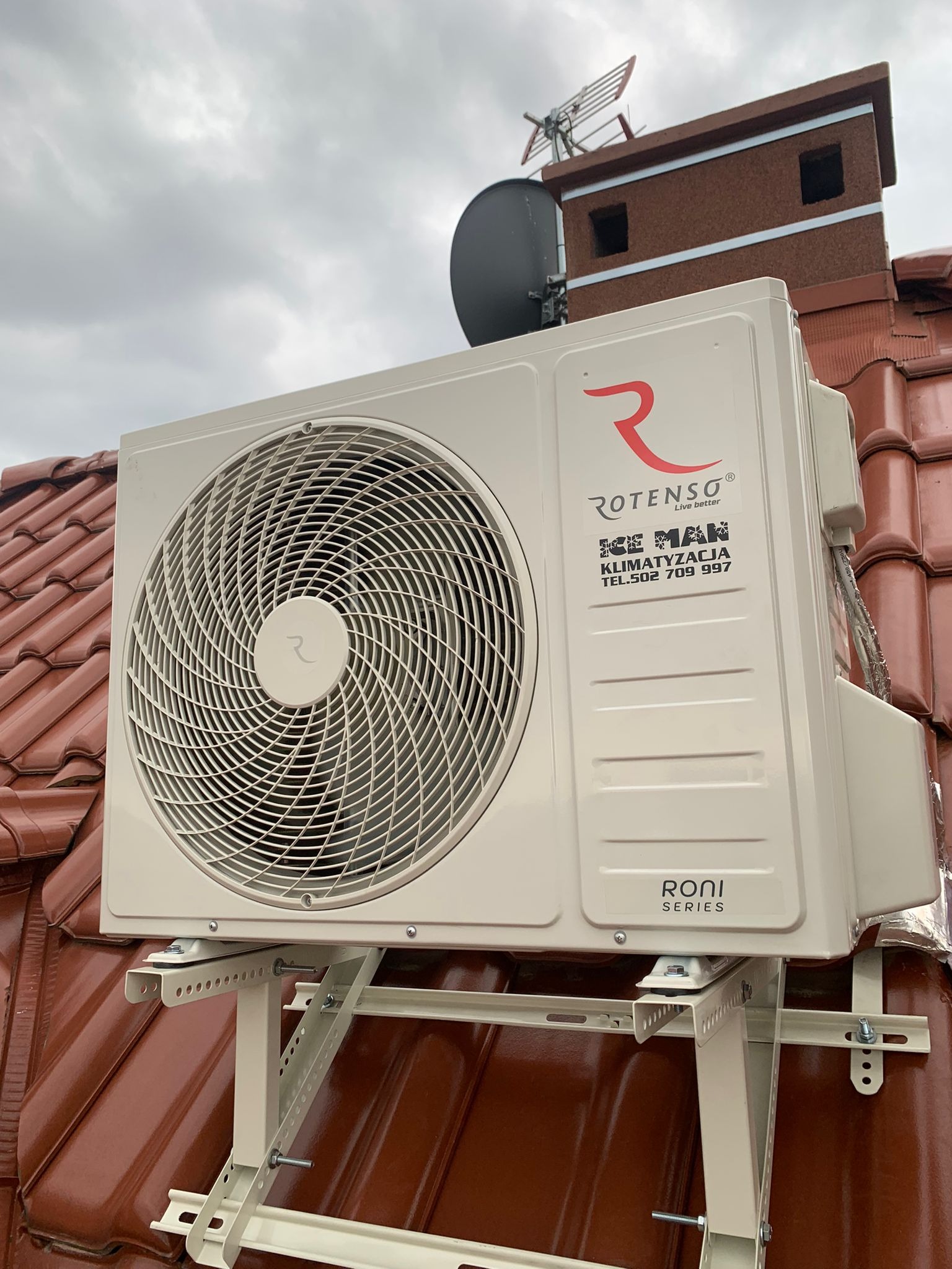 Zewnętrzna jednostka klimatyzacji Rotenso zamontowana na wspornikach na dachu z czerwonej dachówki, widoczny komin i antena satelitarna na tle pochmurnego nieba.