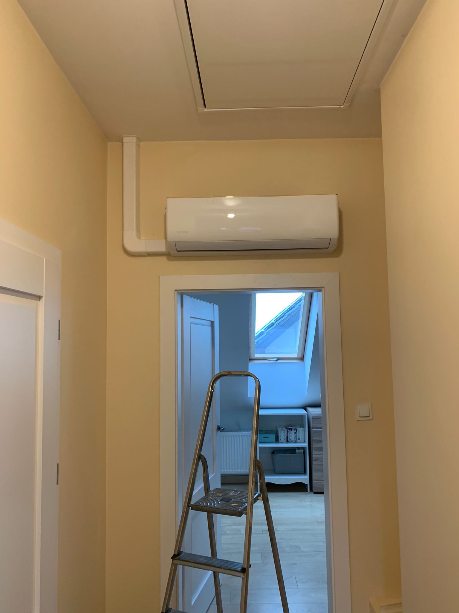 Biały klimatyzator ścienny zamontowany w korytarzu domu, widoczna instalacja w białej listwie, drabina w tle, otwarte drzwi do pokoju z oknem dachowym.