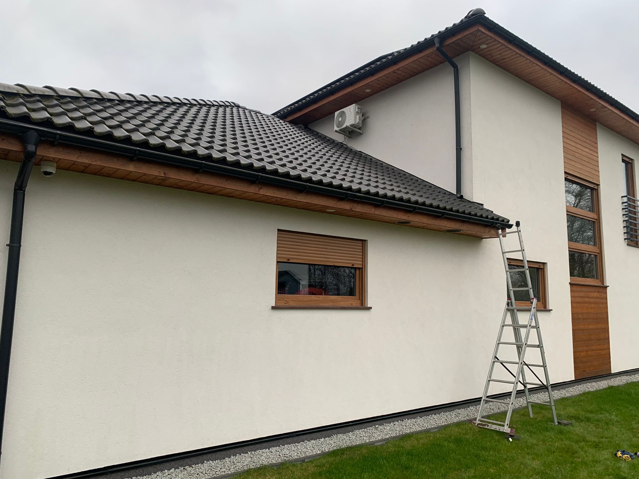 Biały dom jednorodzinny z zamontowaną klimatyzacją na elewacji, widoczna drabina oparta o ścianę, okna z roletami zewnętrznymi.