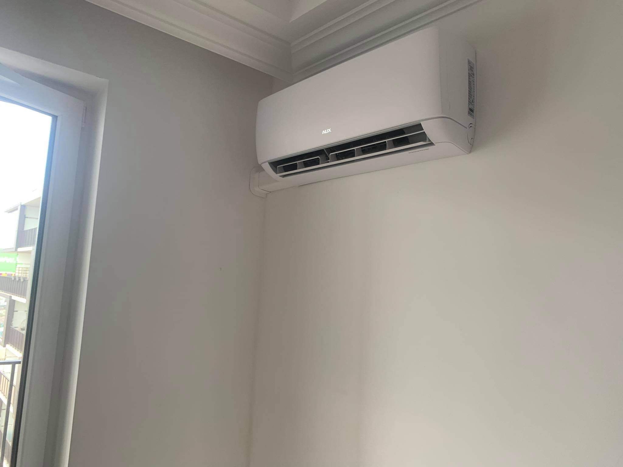 Biała jednostka klimatyzacji marki AUX zamontowana na ścianie w jasnym pomieszczeniu, widok z boku z oknem po lewej stronie.