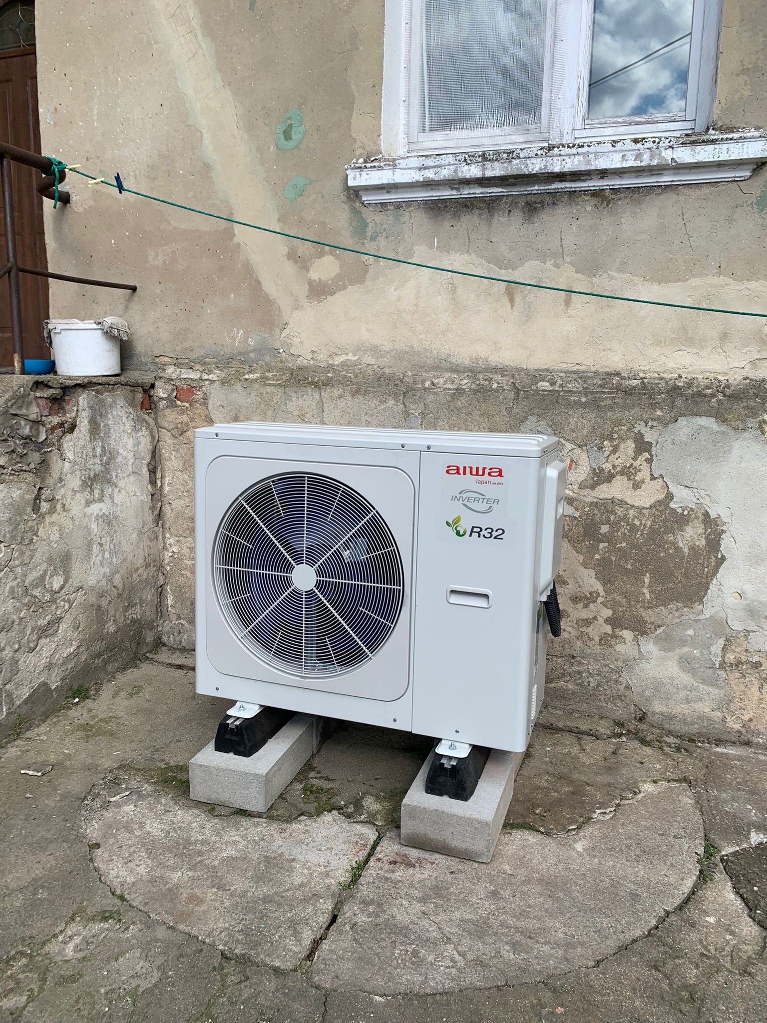 Zewnętrzna jednostka klimatyzacji Aiwa Inverter R32 ustawiona na betonowych bloczkach przed starą, otynkowaną ścianą budynku z oknem i sznurkiem na pranie.