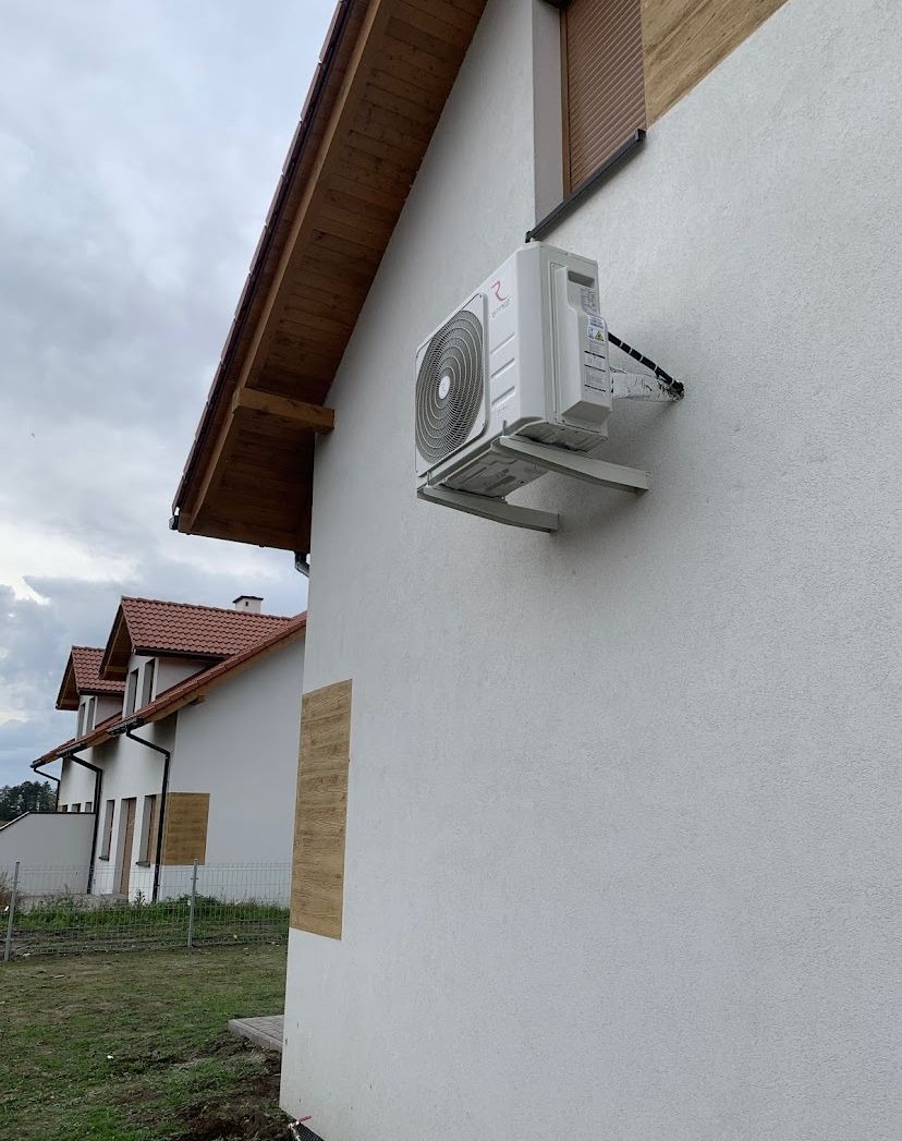 Biała jednostka zewnętrzna klimatyzatora zamontowana na białej ścianie domu jednorodzinnego z drewnianymi elementami wykończeniowymi dachu i okiennicami. Widoczne inne domy w tle.