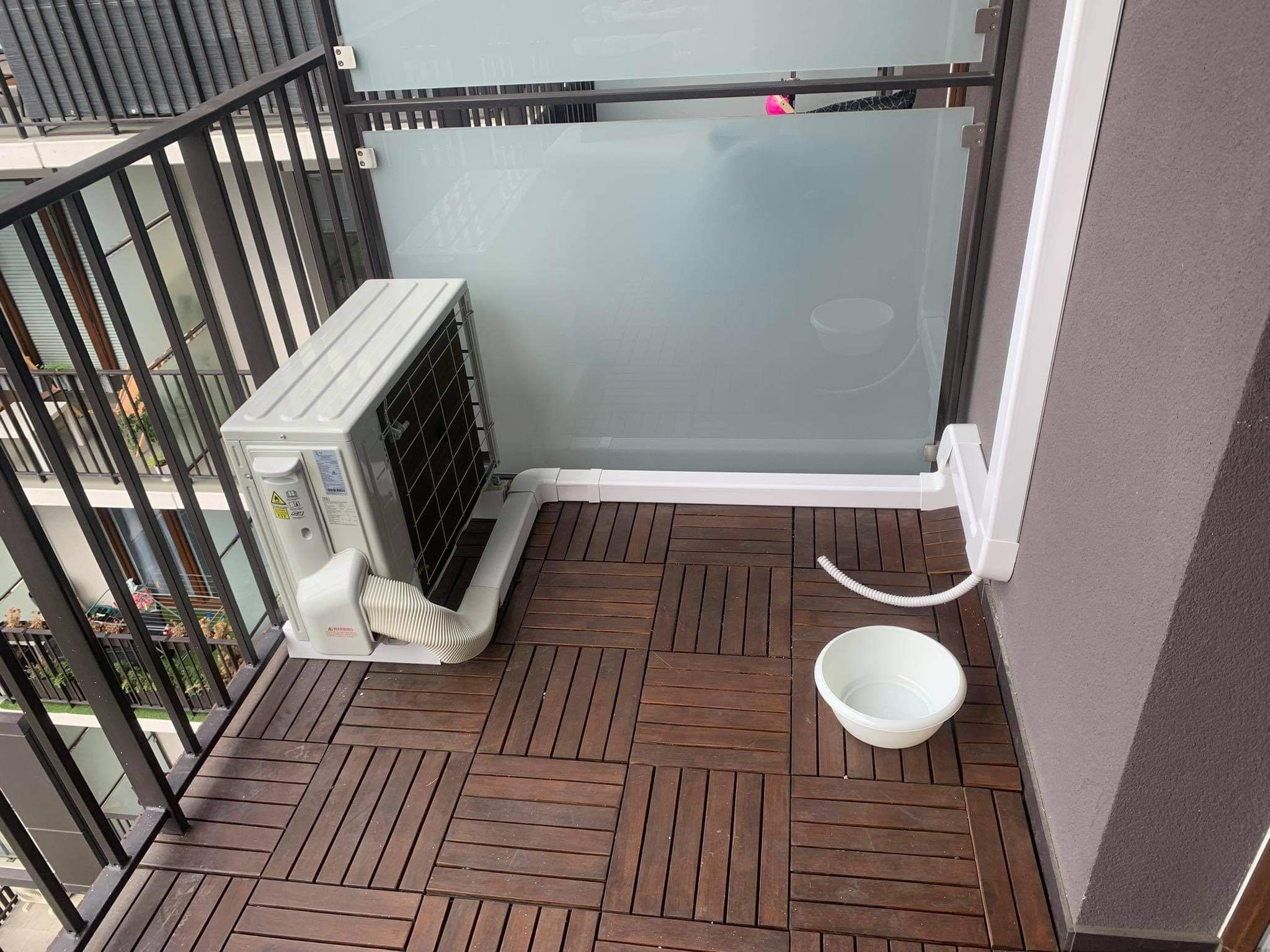 Jednostka zewnętrzna klimatyzacji na balkonie z drewnianą podłogą, białe rury odprowadzające skropliny i miska zbierająca wodę.