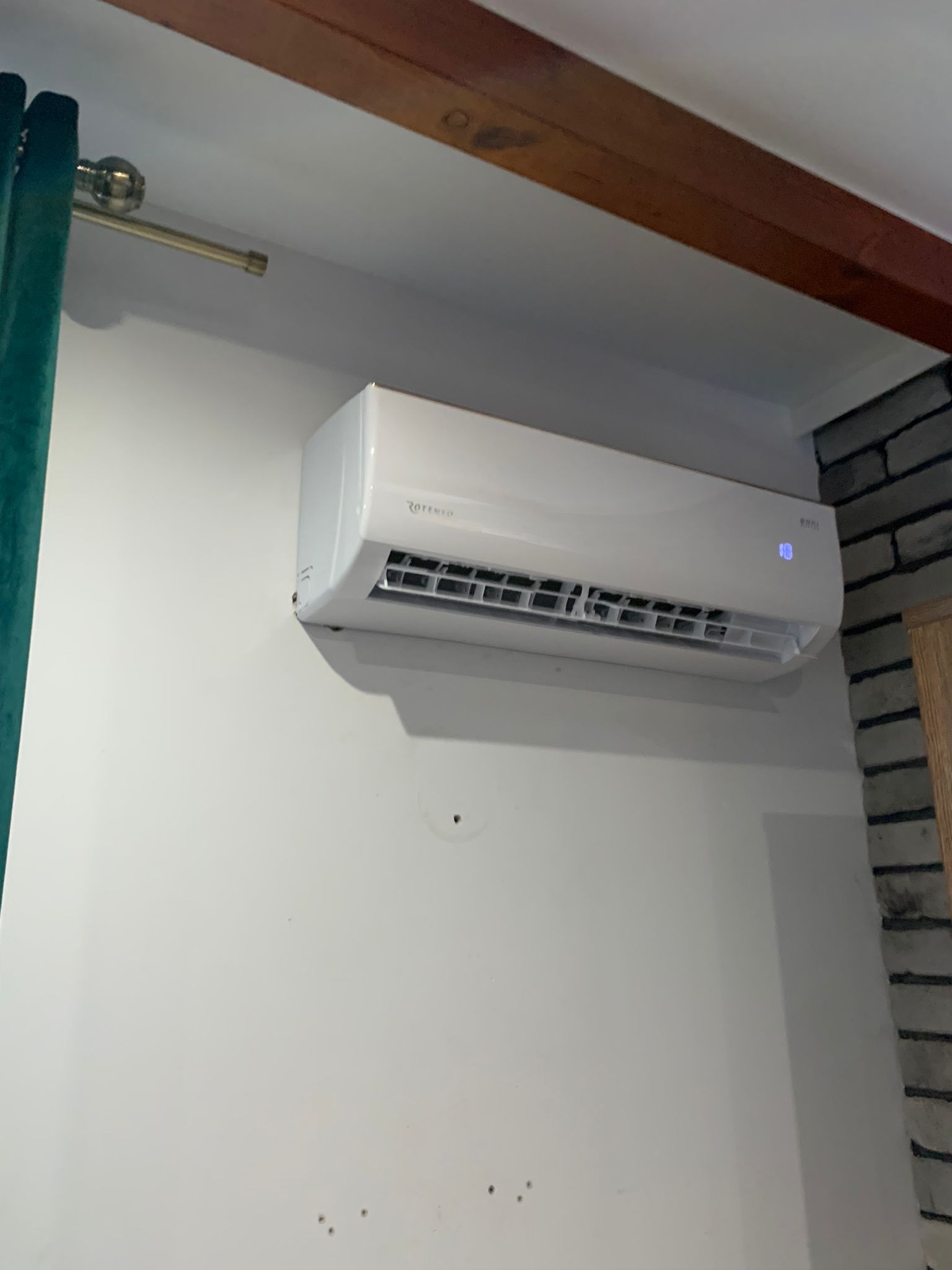 Biały klimatyzator ROTENSO zamontowany na białej ścianie w pomieszczeniu z drewnianą belką sufitową i fragmentem ściany z cegły.