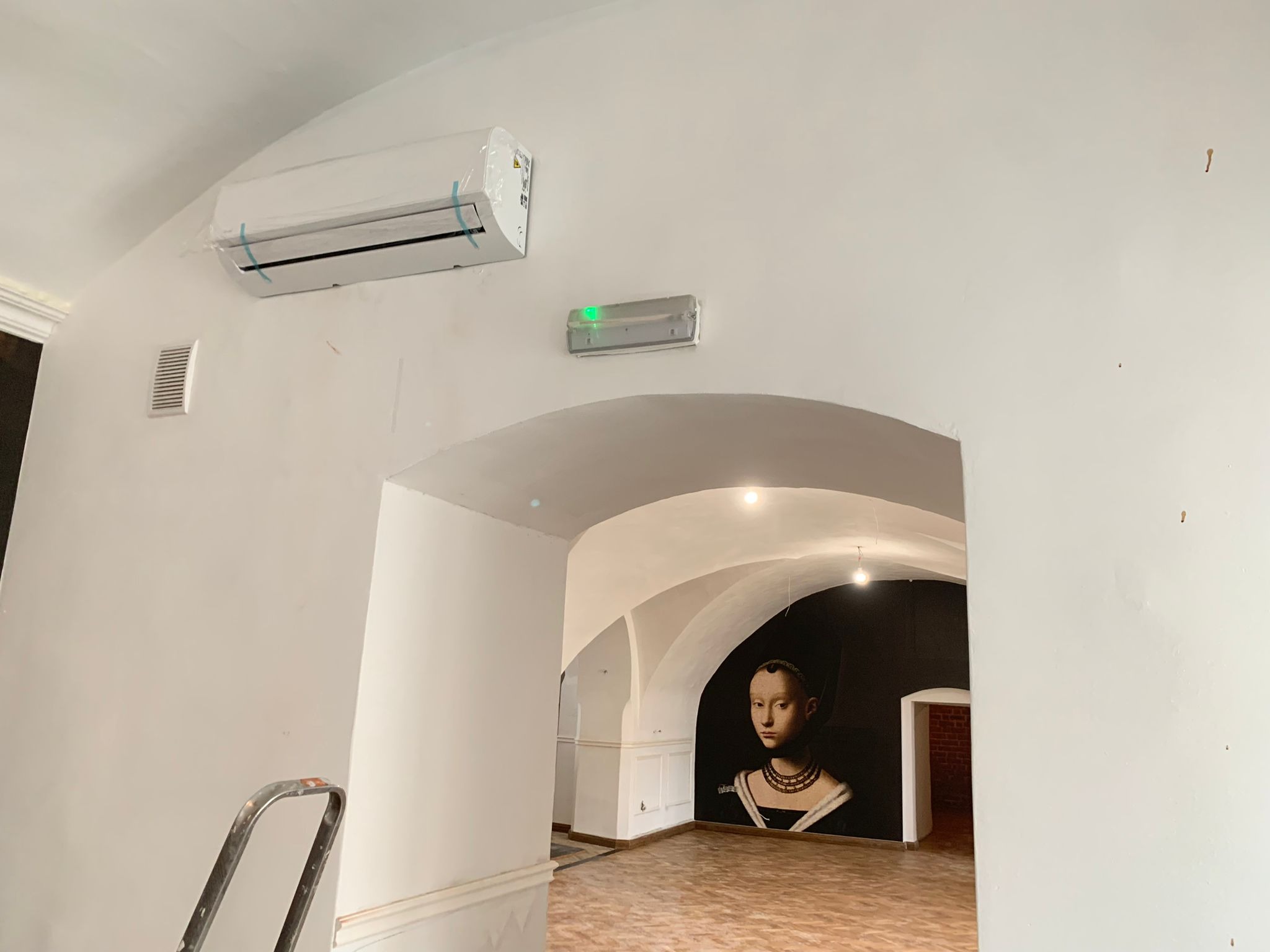 Nowa klimatyzacja ścienna w folii ochronnej, zamontowana w pomieszczeniu z łukowatym sufitem i dekoracyjnym malowidłem ściennym, widoczna drabina.