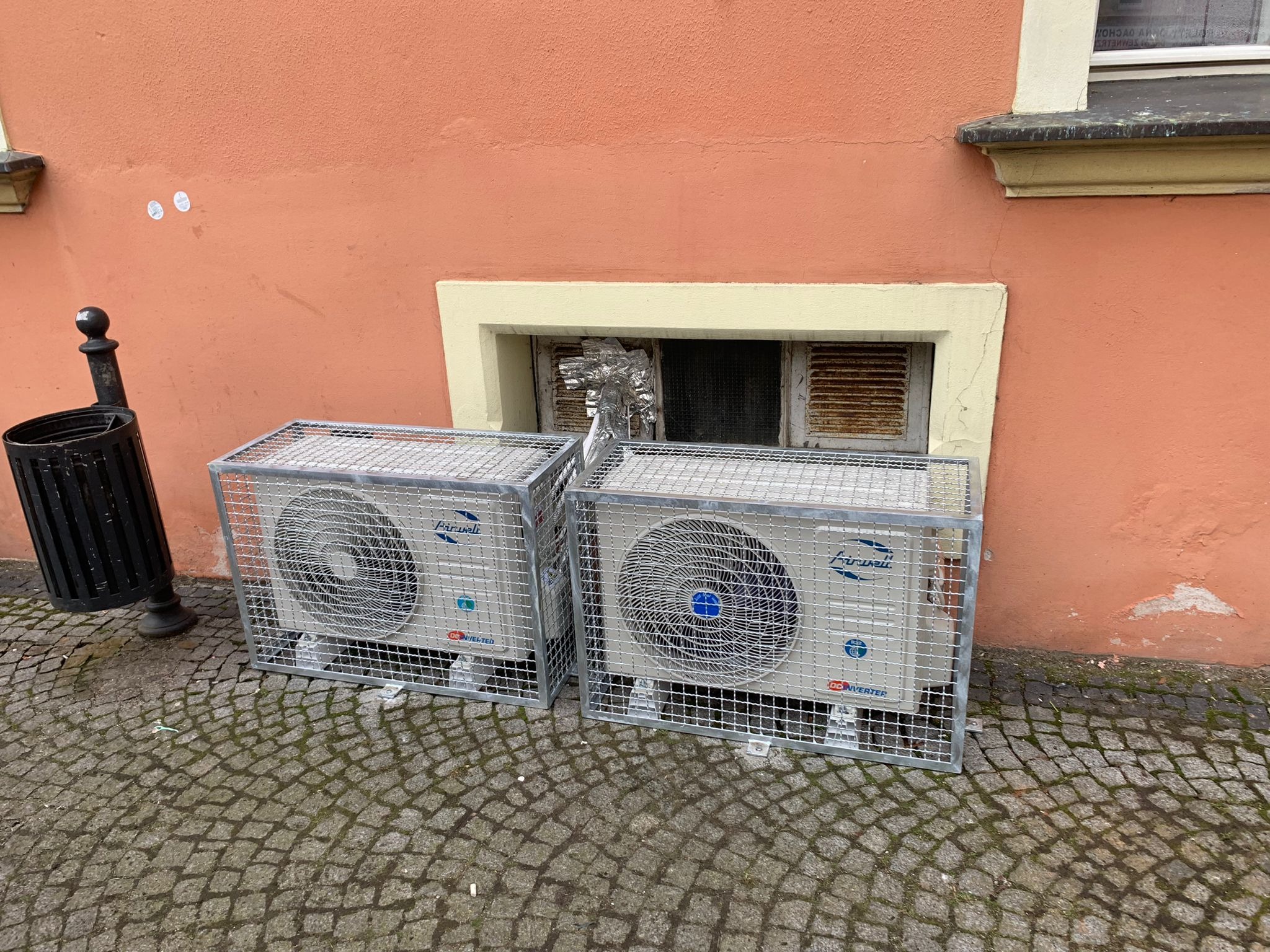 Dwa zewnętrzne moduły klimatyzacji marki Firwall, zabezpieczone metalowymi klatkami, stoją na brukowanym chodniku przed budynkiem z pomarańczową ścianą i małym, zamurowanym oknem zabezpieczonym folią.