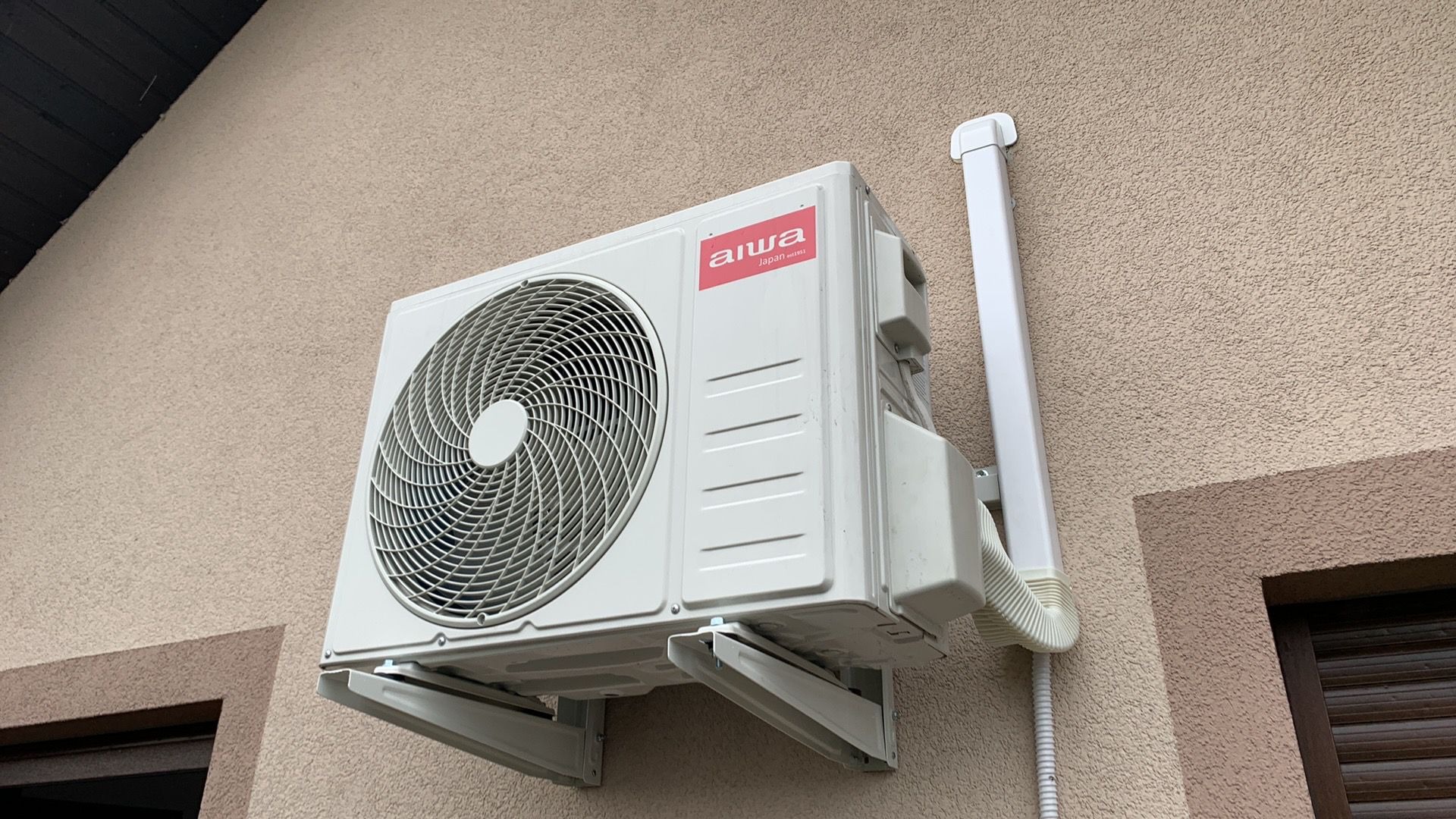 Zewnętrzna jednostka klimatyzacji Aiwa zamontowana na beżowej elewacji budynku, widoczna rura odprowadzająca skropliny, zarys okna z brązowymi okiennicami w tle.
