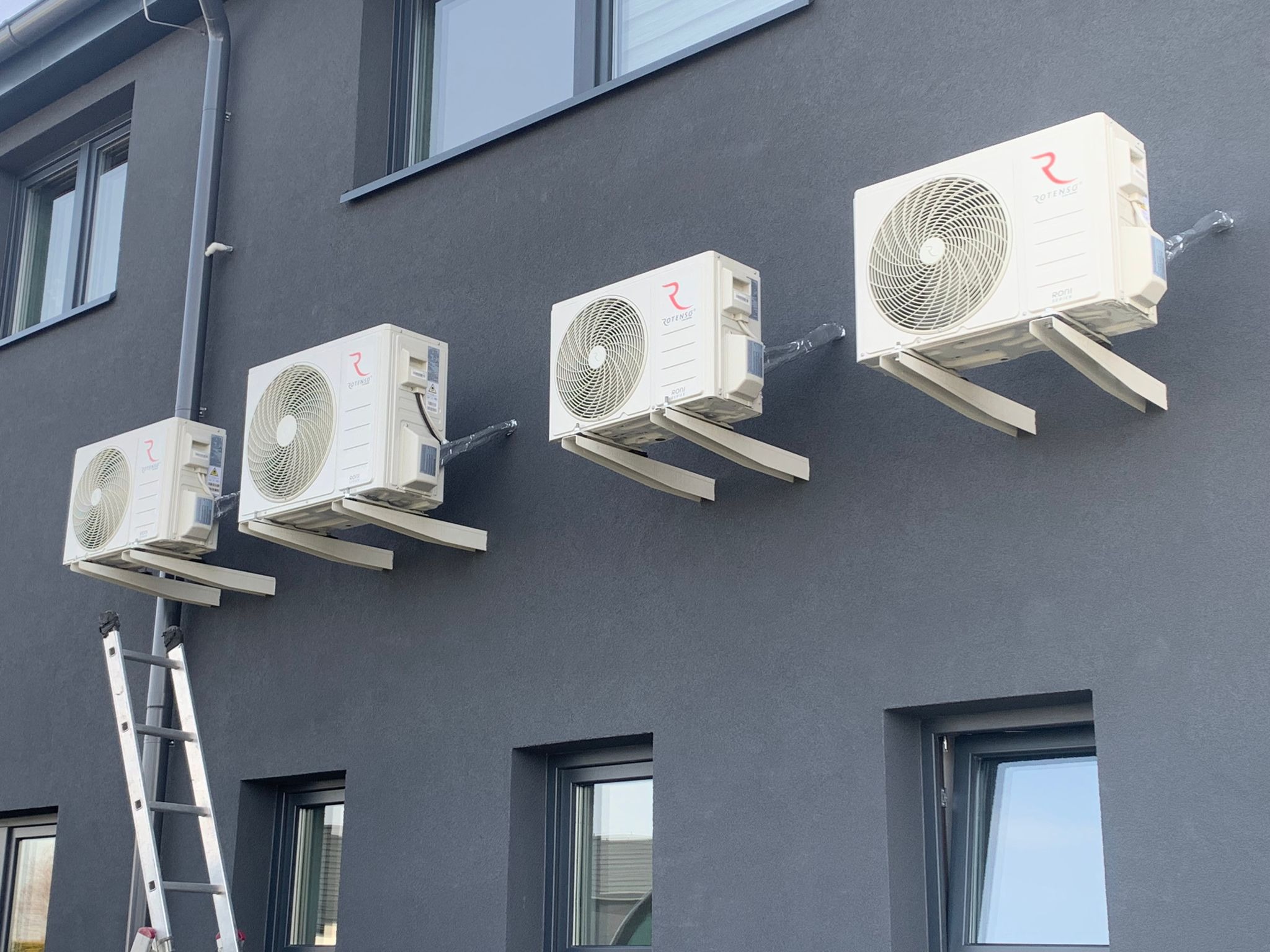 Cztery klimatyzatory Rotenso zamontowane na szarej ścianie budynku, drabina oparta o ścianę, widoczne okna.