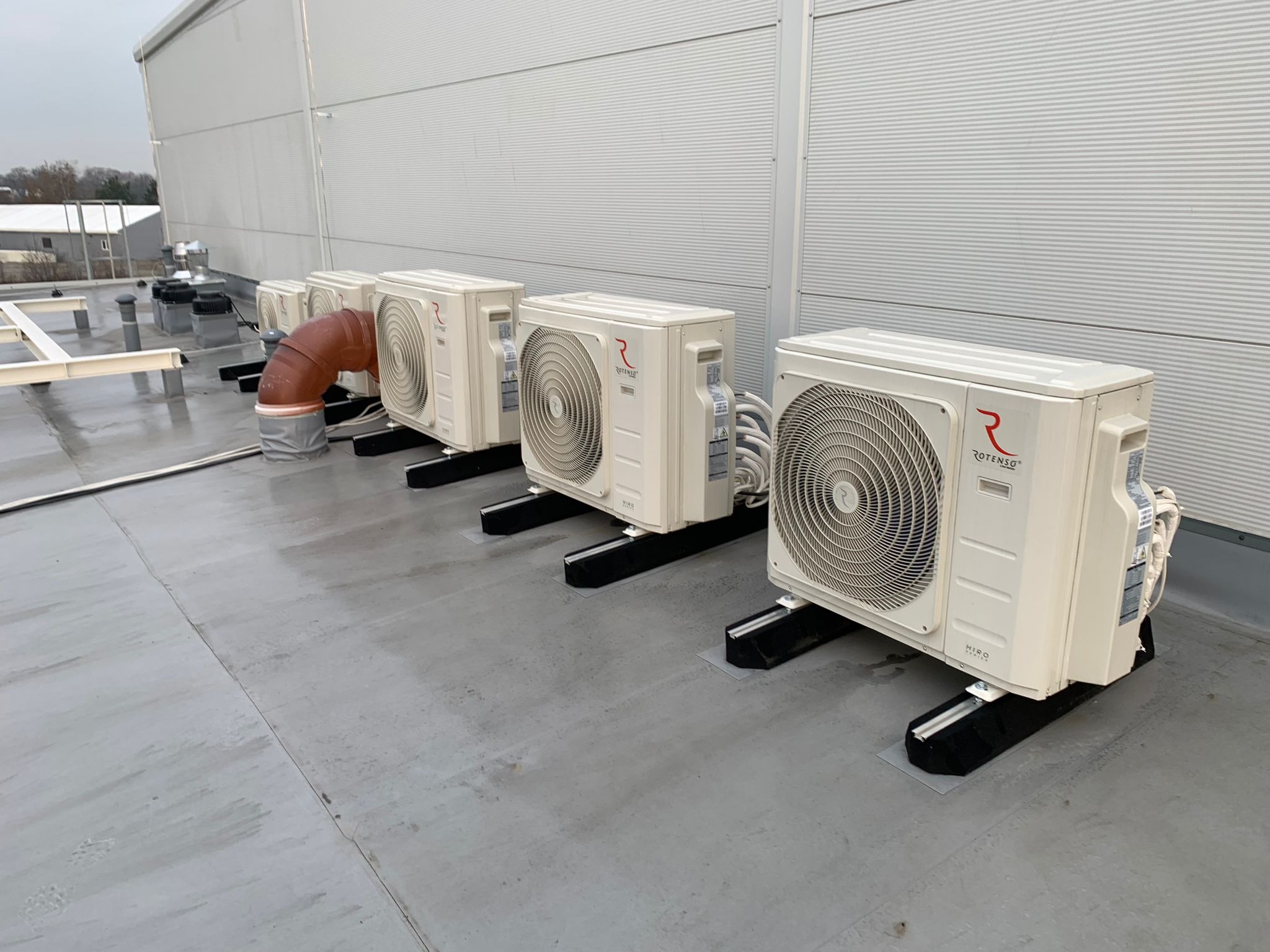 Rząd zewnętrznych jednostek klimatyzacyjnych Rotenso na dachu budynku przemysłowego, połączonych brązową rurą wentylacyjną, widoczne czarne podstawy tłumiące drgania.
