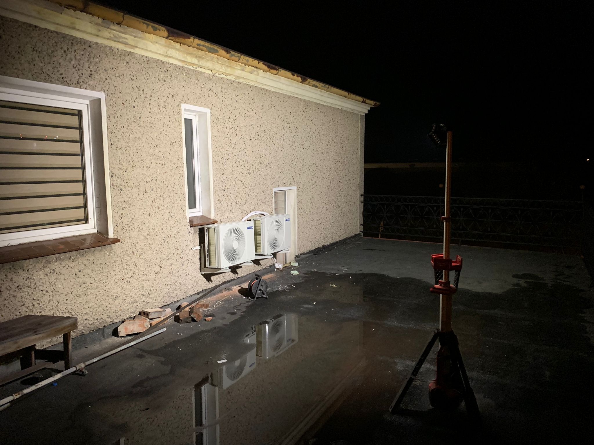 Dwa białe klimatyzatory na ścianie budynku z fakturą baranka, widoczne odbicia w kałużach na dachu, lampa budowlana oświetla teren, cegły i narzędzia obok klimatyzatorów.