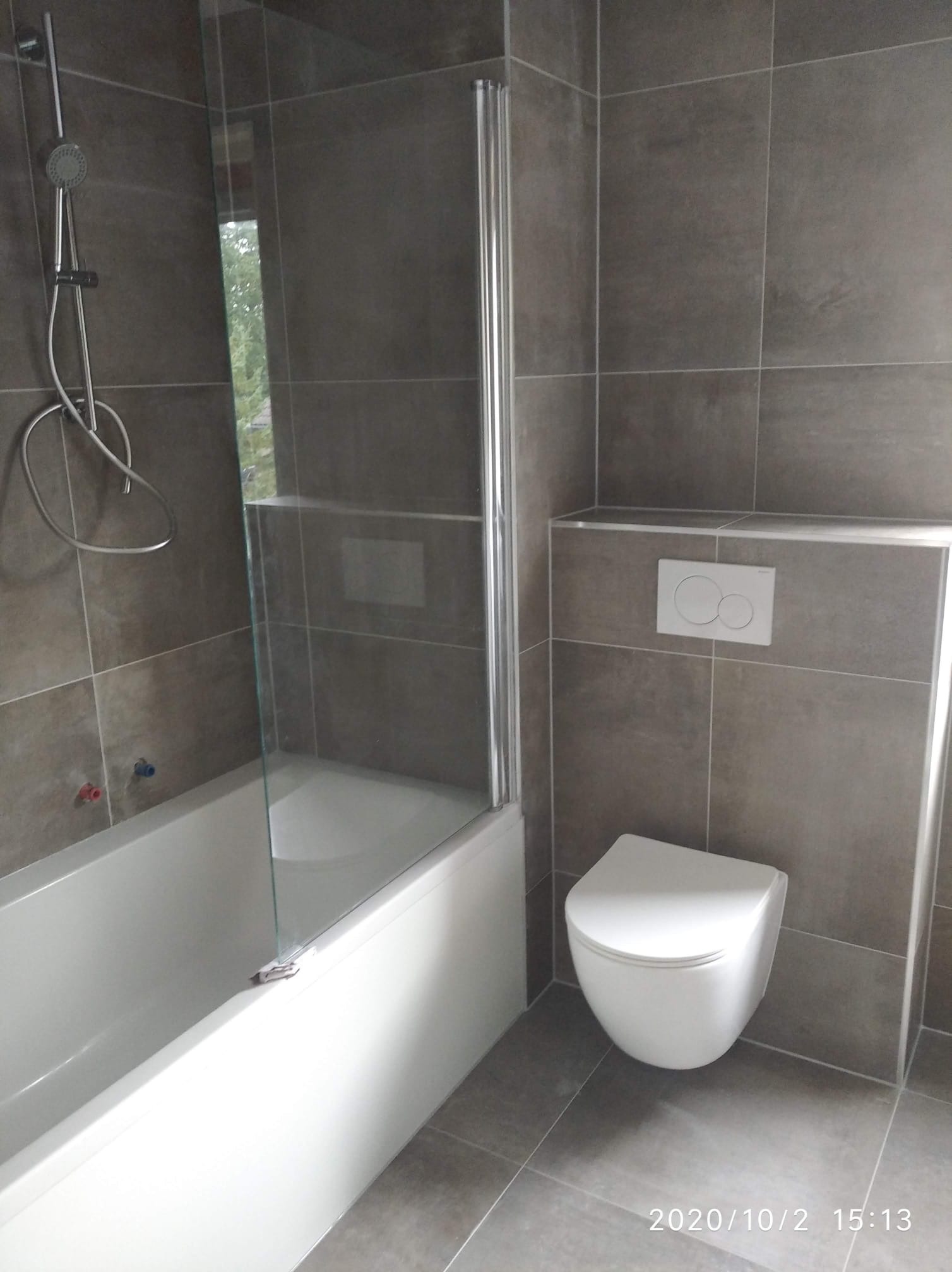 Nowoczesna łazienka z szarymi, kwadratowymi płytkami, białą wanną z szklanym parawanem, wiszącą miską WC i chromowaną baterią prysznicową.