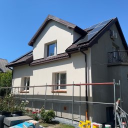 SD-TERM Dominik Skliniorz - Dom z panelami słonecznymi w trakcie prac elewacyjnych, widoczne rusztowania i farby. Część ściany otynkowana na beżowo, reszta w szarym podkładzie.