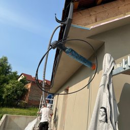 SD-TERM Dominik Skliniorz - Prace elewacyjne w toku: robotnicy na rusztowaniu, widoczne ocieplenie dachu styropianem, szare wykończenie elewacji, kabel wentylacyjny i bluza robocza na drabinie.