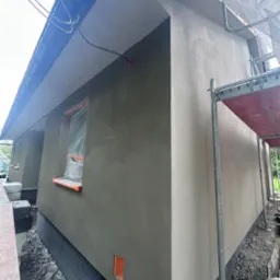 Świeżo otynkowana, szara elewacja domu jednorodzinnego z widocznymi rusztowaniami i zabezpieczonym oknem folią. Widoczna siatka drenażowa przy fundamencie.