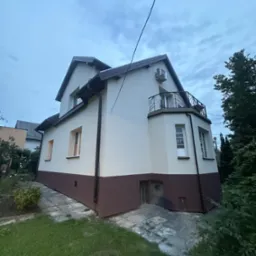 Dwukondygnacyjny dom z balkonem, świeżo po malowaniu elewacji w tonacji beżowo-brązowej. Widoczny fragment ogrodu i niebo z chmurami.