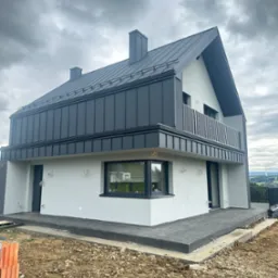 Nowoczesny dom z szarą elewacją i metalowym dachem na tle pochmurnego nieba. Widoczny balkon z metalową balustradą i częściowo wykończony teren wokół budynku.