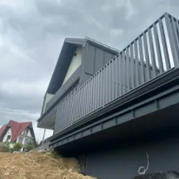 Nowoczesna elewacja domu z szarym wykończeniem i metalową balustradą. W tle widoczny inny budynek z czerwoną dachówką. Pochmurne niebo.