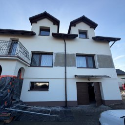 SD-TERM Dominik Skliniorz - Dwupiętrowy dom z białymi ścianami i brązowymi akcentami, w trakcie prac elewacyjnych. Widoczne rusztowania i zabezpieczenia. Fragmenty ścian przygotowane do malowania.