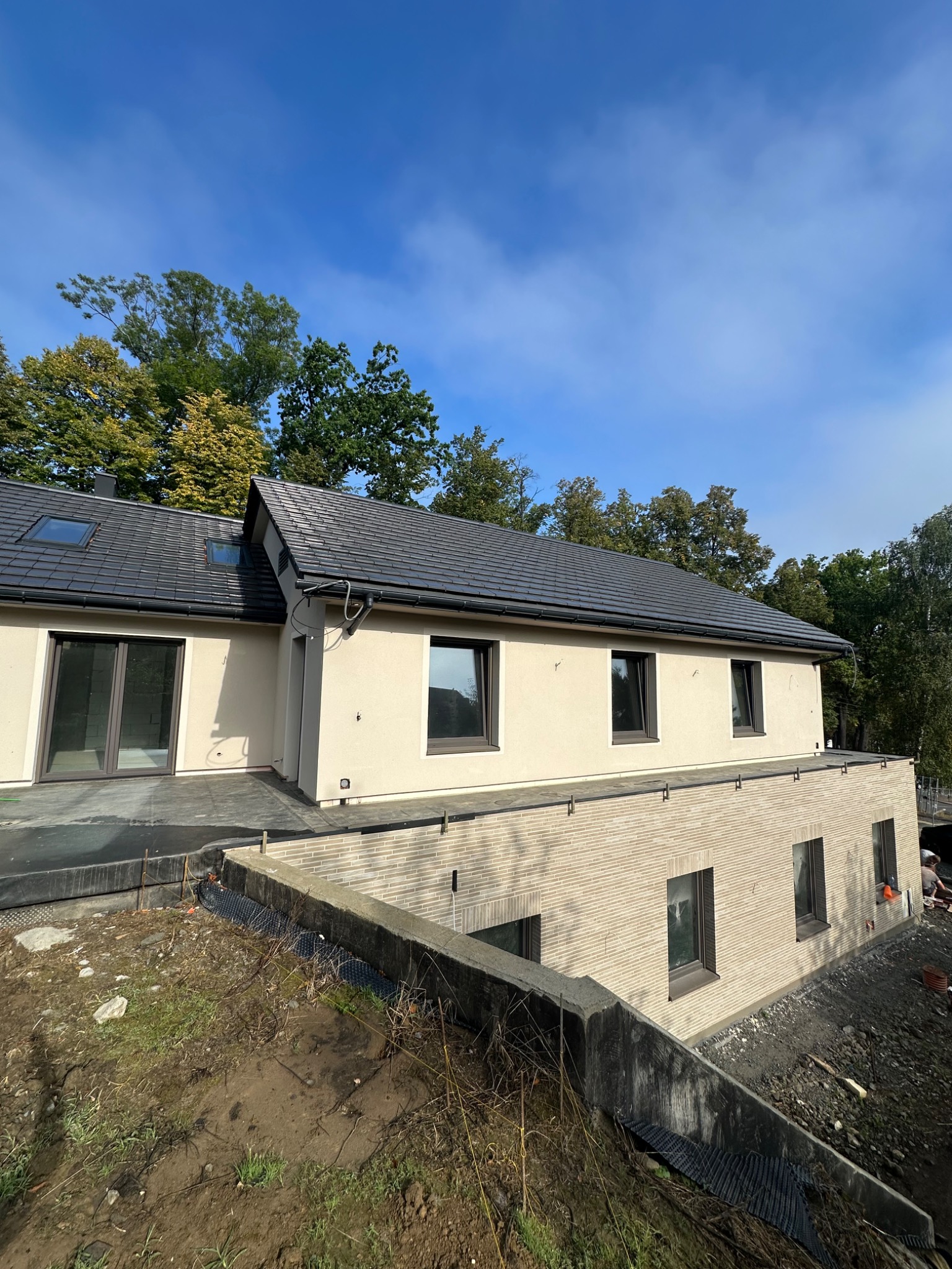 Nowoczesny dom z beżową elewacją i ciemną dachówką, widoczny z perspektywy gruntu. Częściowo wykończony teren wokół domu, błękitne niebo w tle.