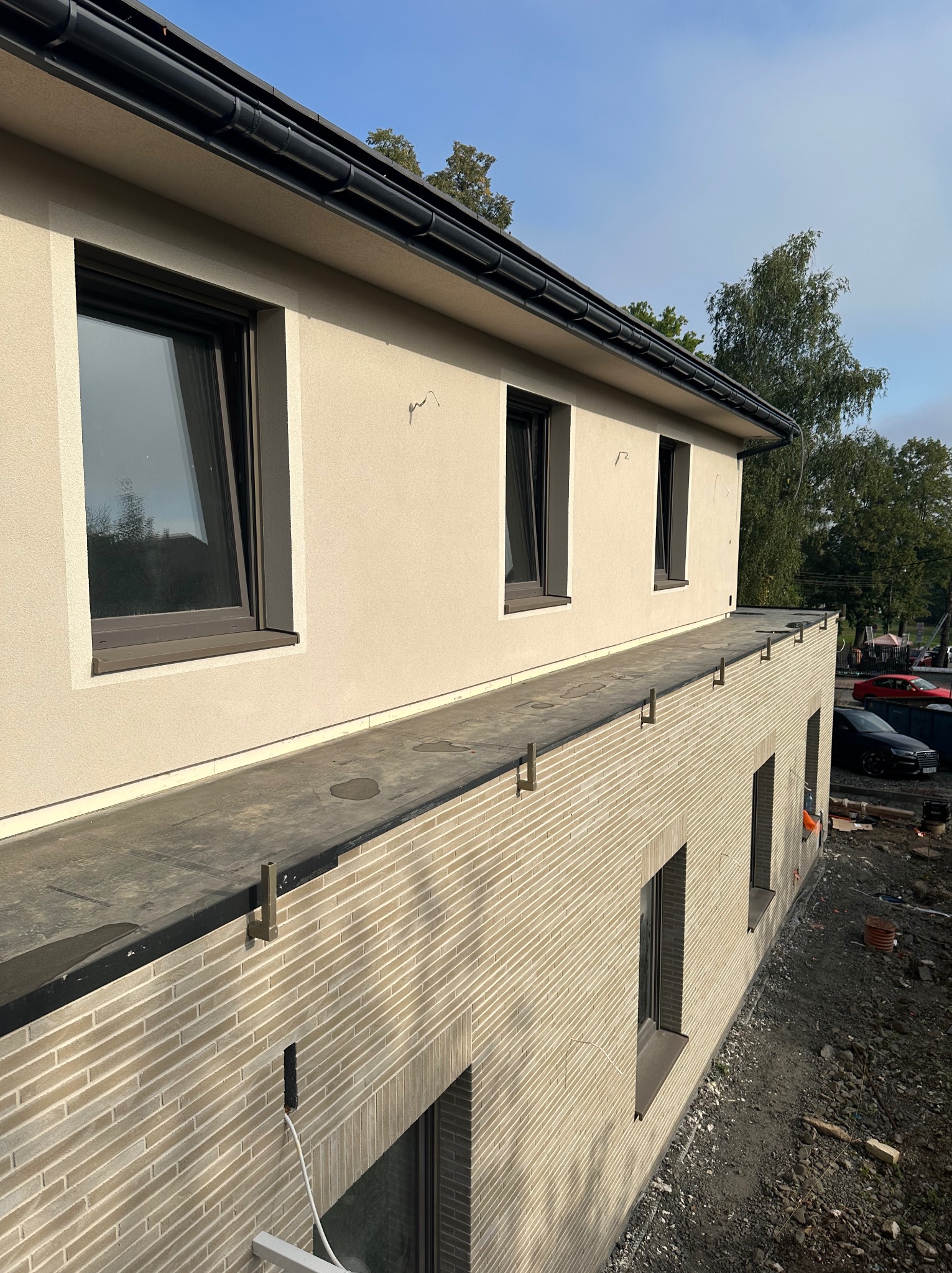 Elewacja dwukondygnacyjnego budynku z beżowym tynkiem i wykończeniem z imitacji cegły. Widoczne okna z ciemnymi ramami, rynny i fragment dachu. Prace wykończeniowe w toku.