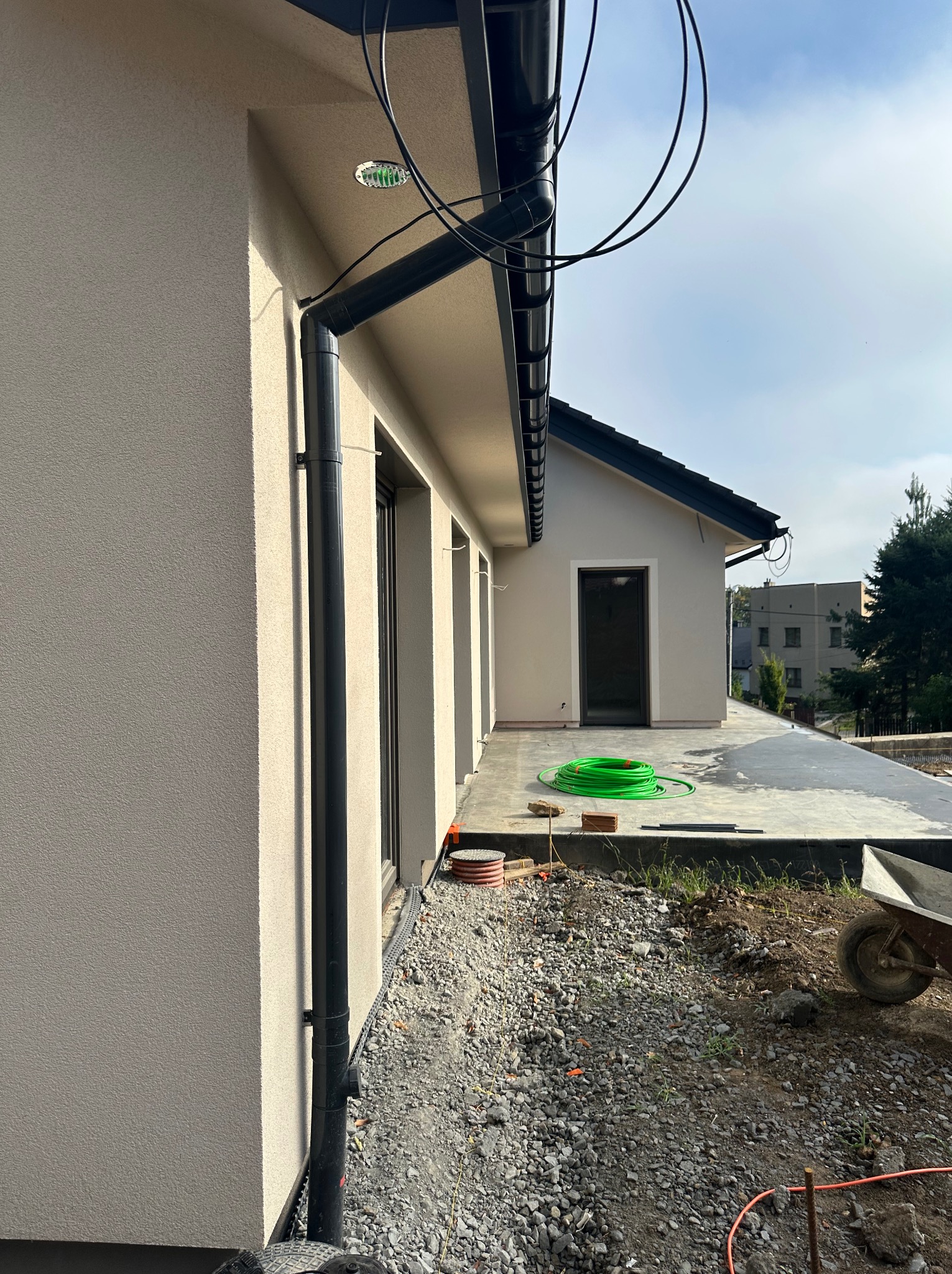 Jasna elewacja domu jednorodzinnego z czarną rynną, widok z boku. Na tarasie zwinięty zielony wąż, w tle zabudowa mieszkalna i drzewa. Drobnoziarnisty tynk.