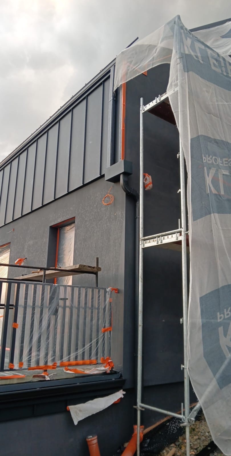 Dom z szarą elewacją, częściowo zakryty rusztowaniem i folią ochronną. Widoczne okna zabezpieczone folią i elementy instalacji. Detal elewacji.
