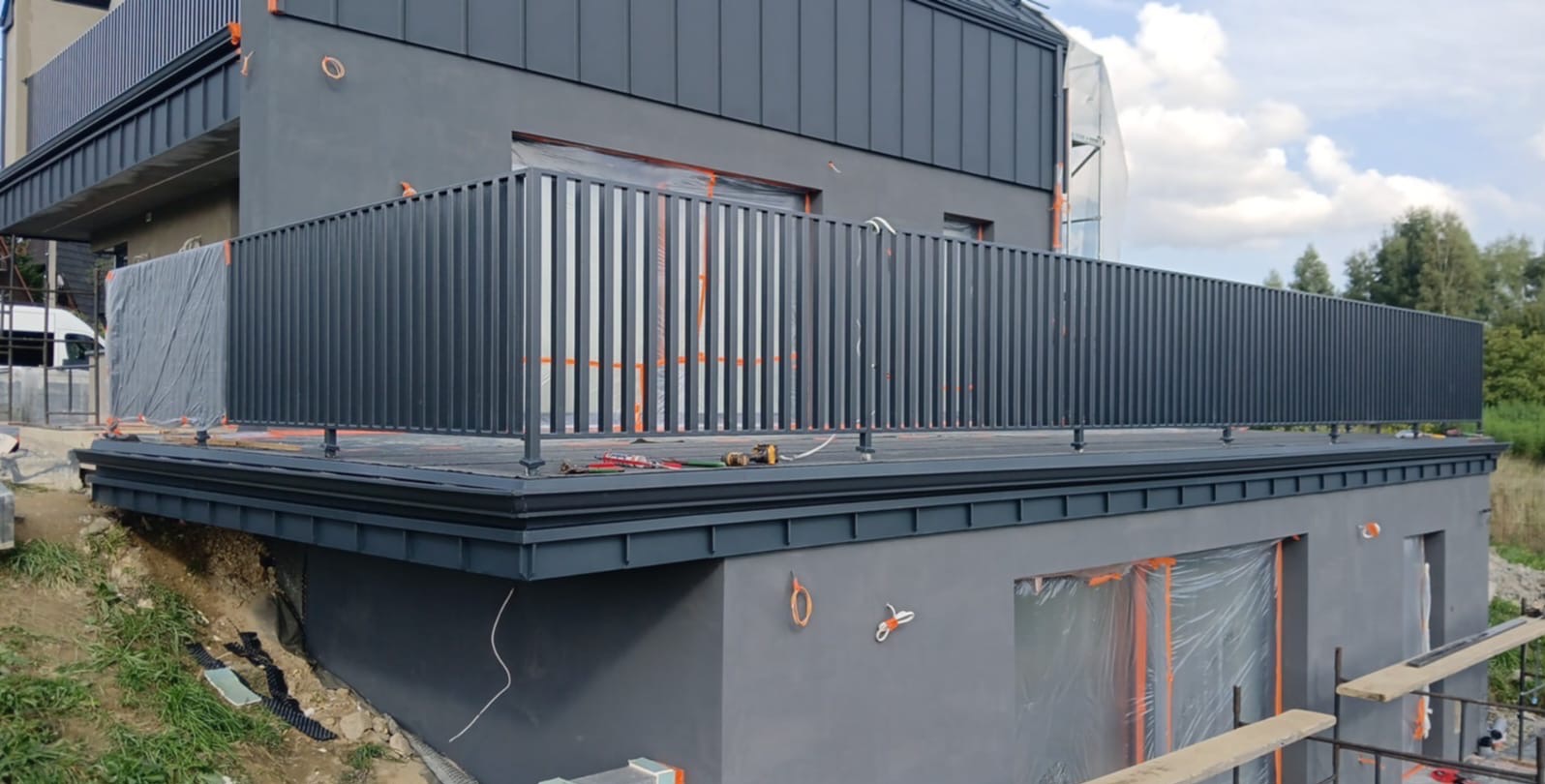 Elewacja domu z szarą, nowoczesną fasadą i balustradą. Okna zabezpieczone folią malarską. Widoczne narzędzia na balkonie.