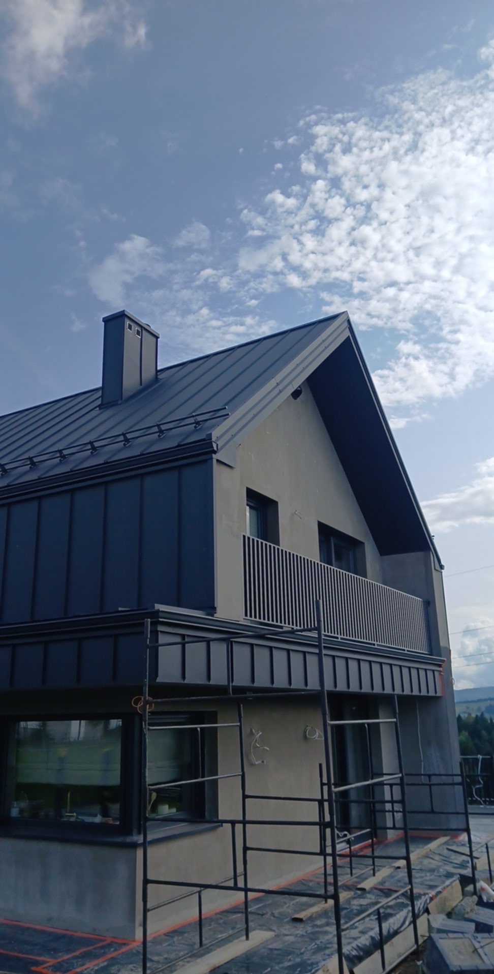 Dom z szarą elewacją i metalowym dachem w trakcie budowy, widoczne rusztowania i elementy wykończeniowe. Ujęcie z dołu, na tle błękitnego nieba.