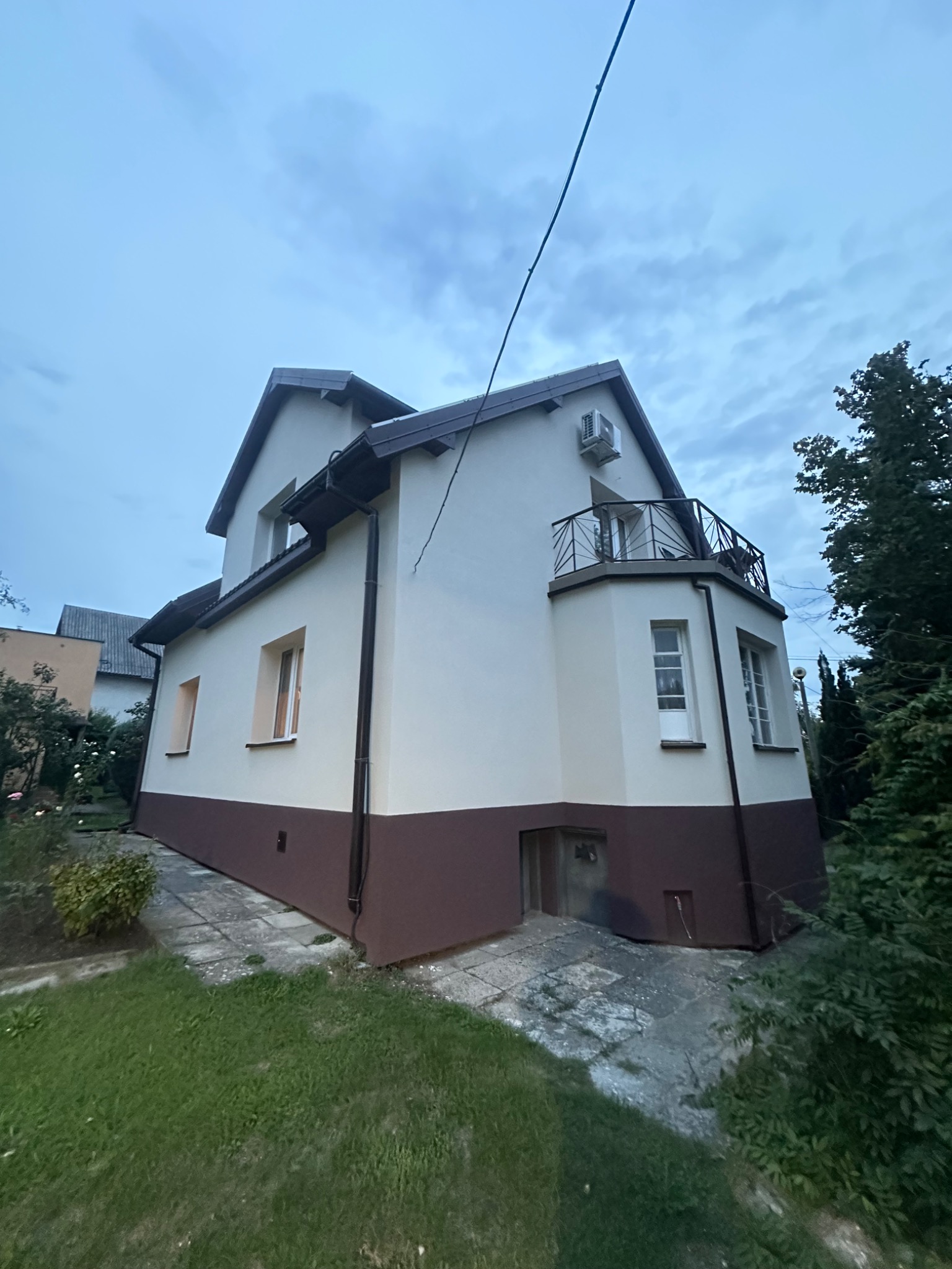 Dwukondygnacyjny dom z balkonem, świeżo po malowaniu elewacji w tonacji beżowo-brązowej. Widoczny fragment ogrodu i niebo z chmurami.