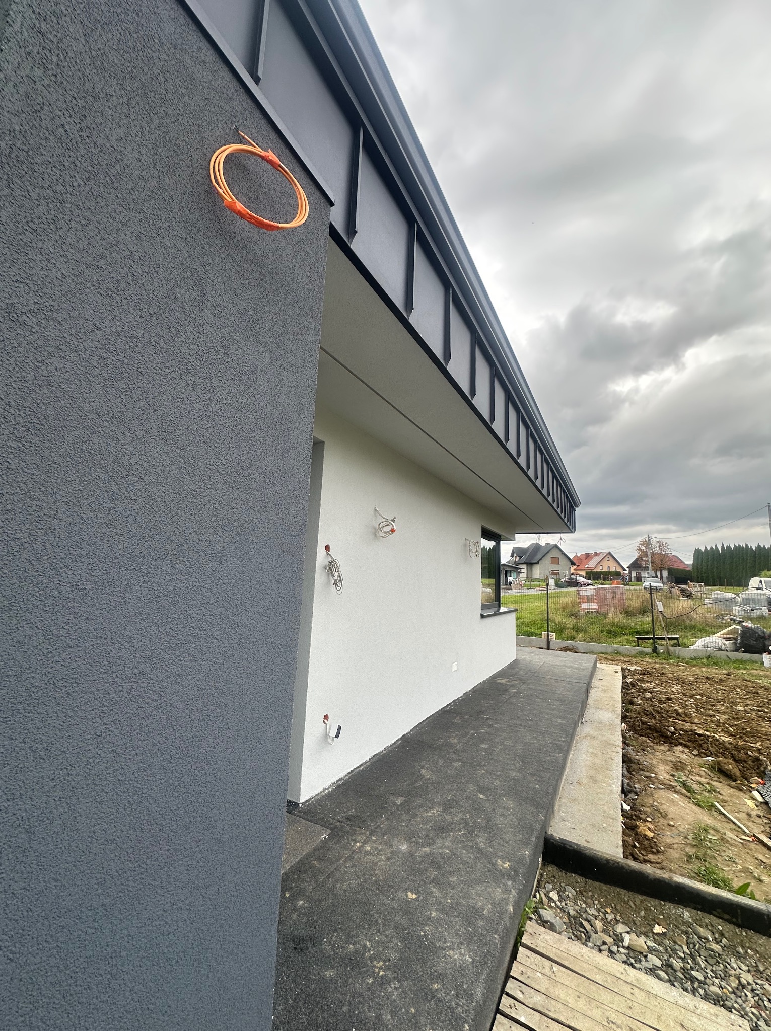 Szara elewacja domu jednorodzinnego z widoczną instalacją elektryczną w pomarańczowej osłonie. W tle budynki mieszkalne i pochmurne niebo.
