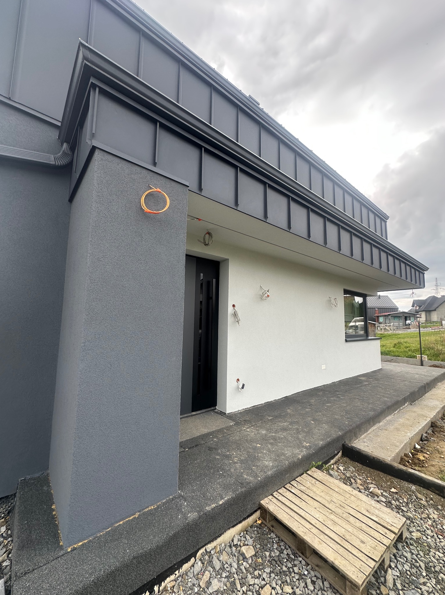 Dwukolorowa elewacja domu w trakcie wykończenia, z widocznymi przewodami instalacji i paletą na podłożu. Szara bryła i biała ściana pod dachem.