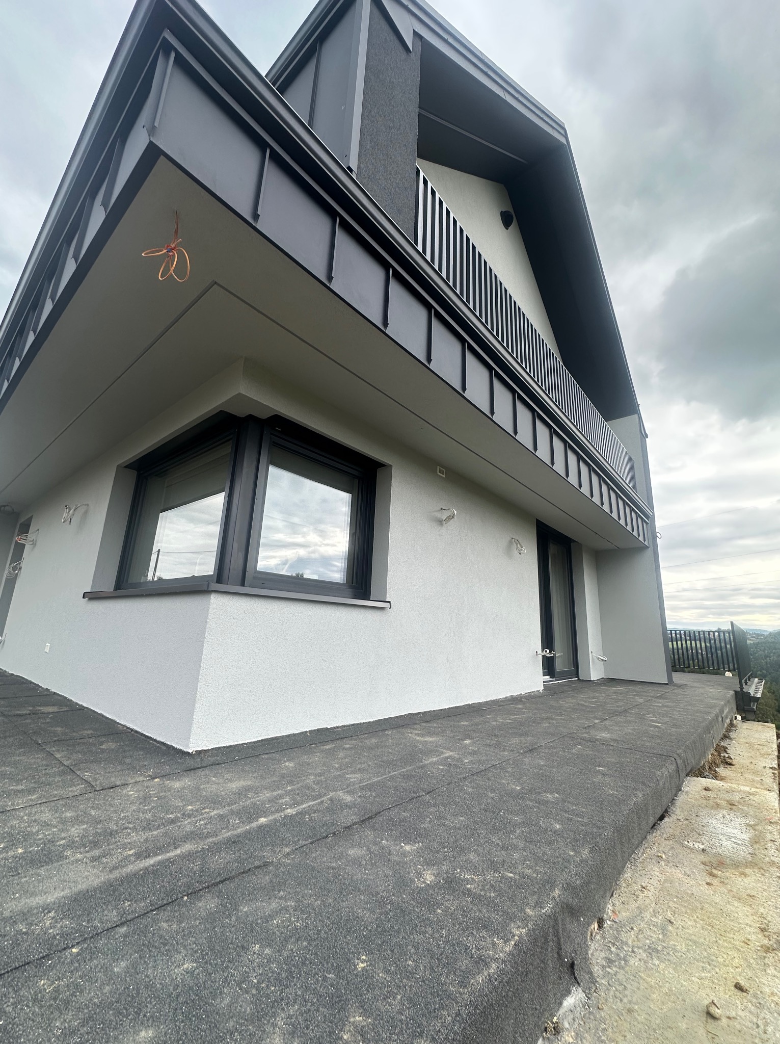 Nowoczesny dom z szarą elewacją i czarnymi detalami, widok z dołu, podkreślający geometrię budynku i fakturę tynku. Widoczne elementy wykończeniowe na balkonie.