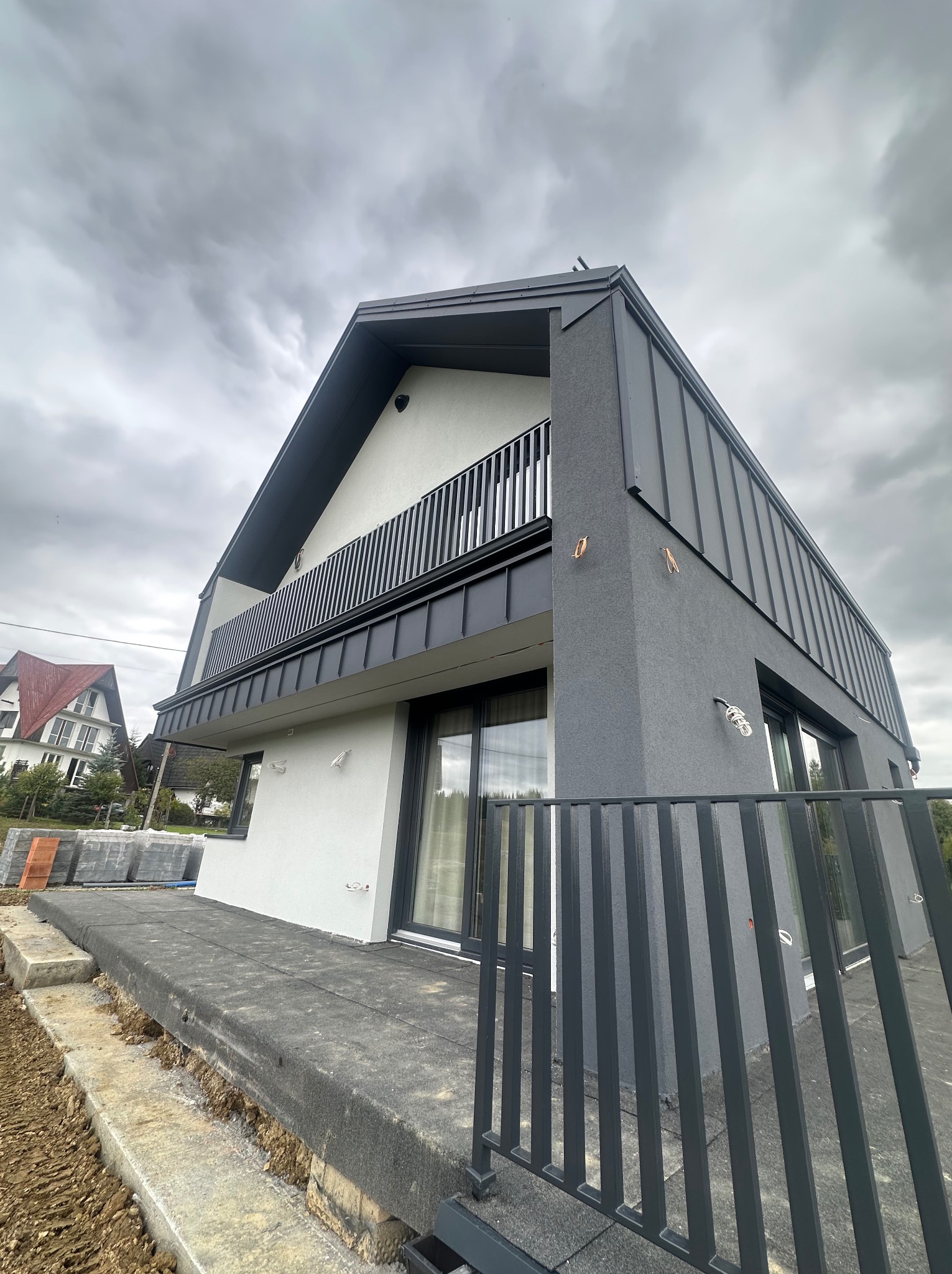 Nowoczesny dom z jasną elewacją i grafitowym wykończeniem dachu oraz balkonu. Ujęcie z dołu podkreśla bryłę budynku na tle pochmurnego nieba.