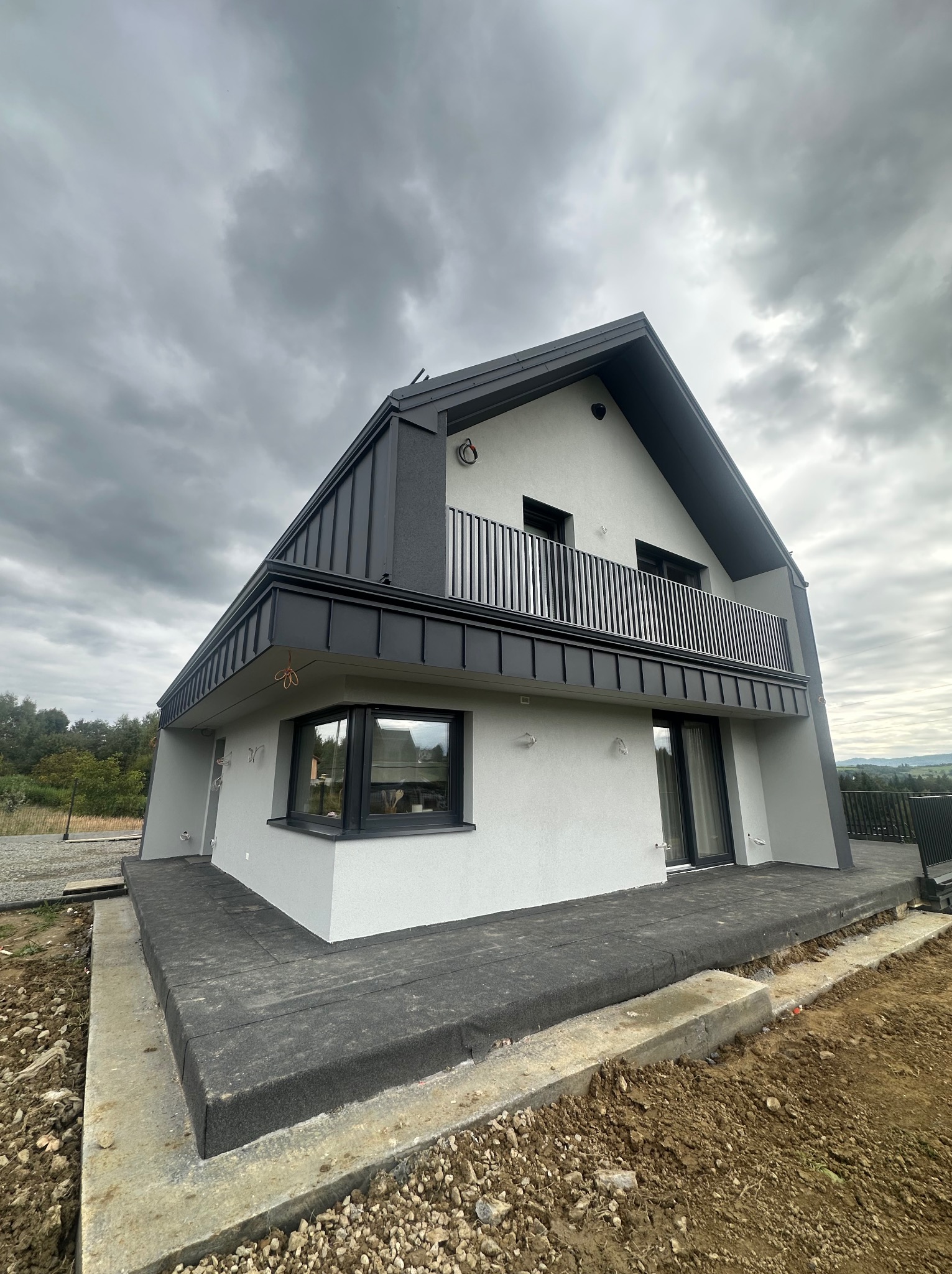 Nowoczesny dom z balkonem i szarą elewacją w trakcie wykończenia, widok z dołu, pochmurne niebo w tle.