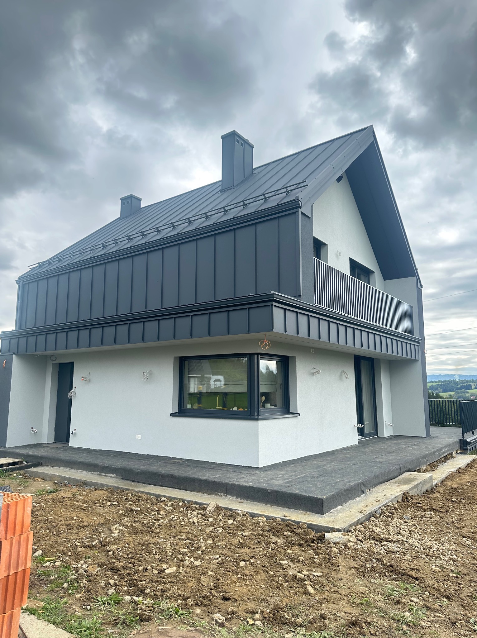 Nowoczesny dom z szarą elewacją i metalowym dachem na tle pochmurnego nieba. Widoczny balkon z metalową balustradą i częściowo wykończony teren wokół budynku.