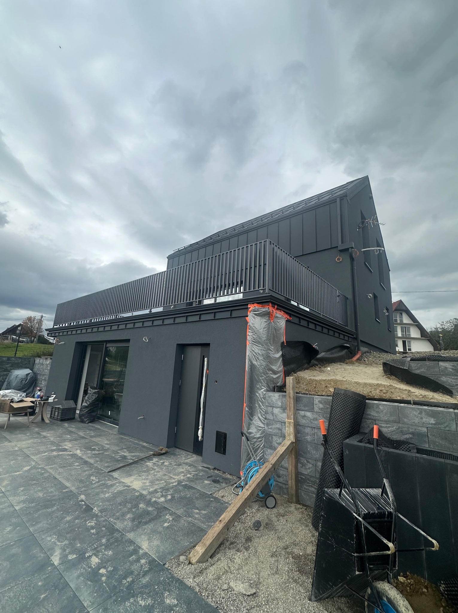 Nowoczesny dom z grafitową elewacją i balkonem, częściowo w trakcie prac wykończeniowych. Widoczne materiały budowlane i narzędzia. Pochmurne niebo w tle.