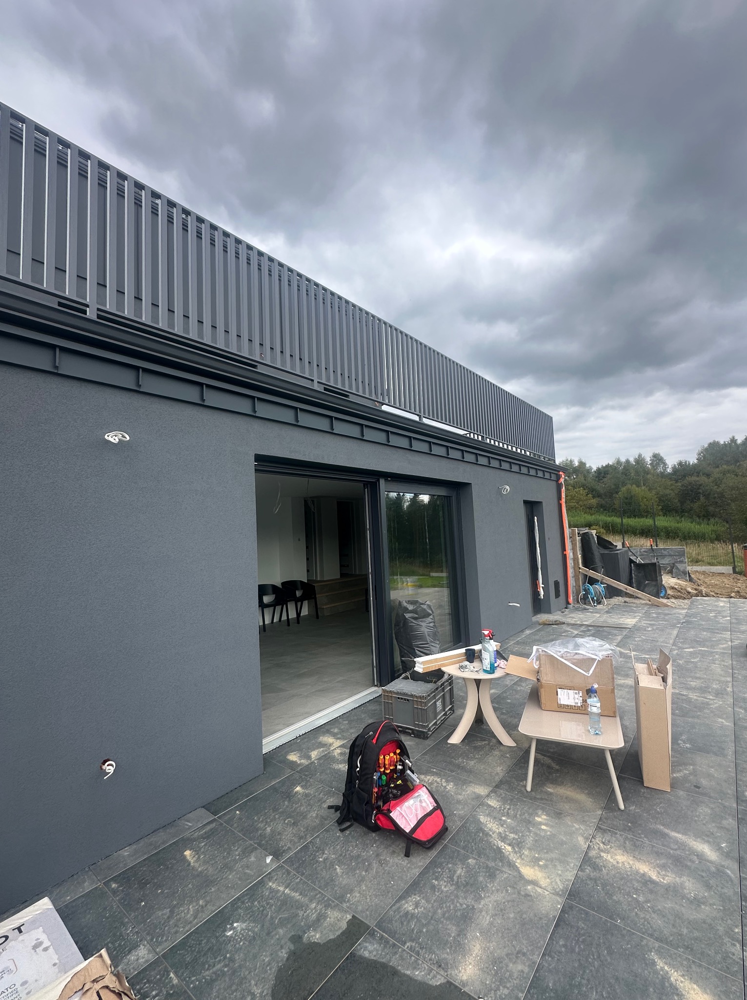 Nowoczesny dom z szarą elewacją i metalowymi balustradami na dachu. Widoczne otwarte przesuwne drzwi i narzędzia pracy na zewnątrz budynku. Pochmurne niebo w tle.