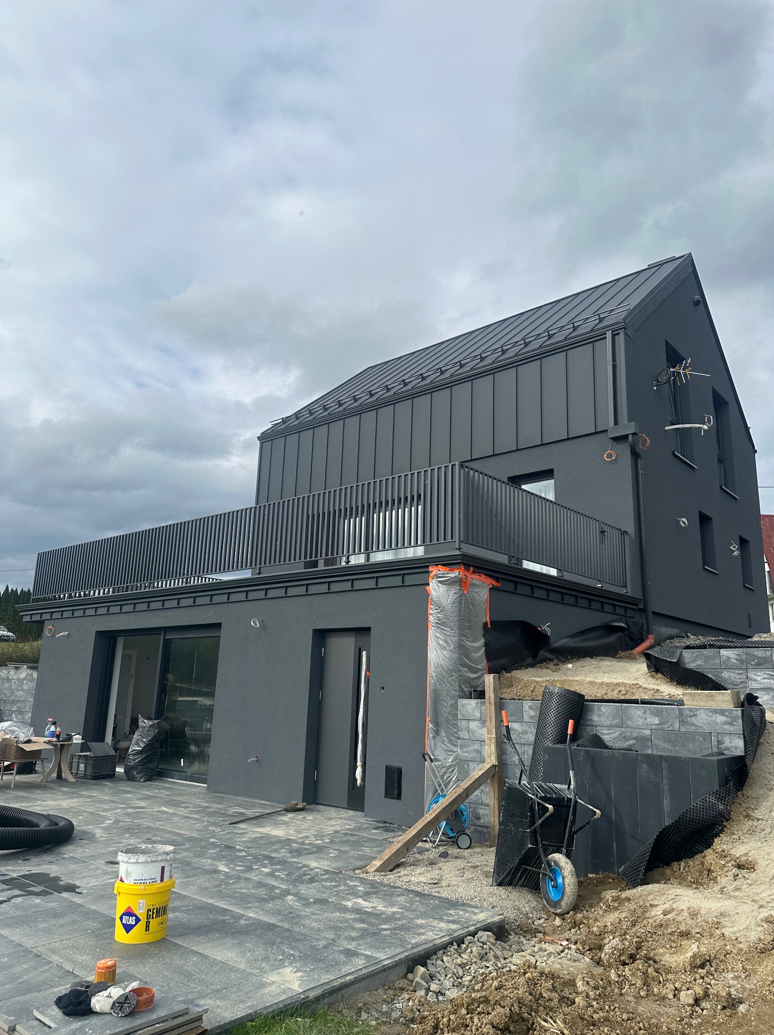 Nowoczesny dom z grafitową elewacją i metalowym dachem na etapie wykończenia. Widoczne narzędzia i materiały budowlane. Uwagę przykuwa spójna kolorystyka i minimalistyczny design.