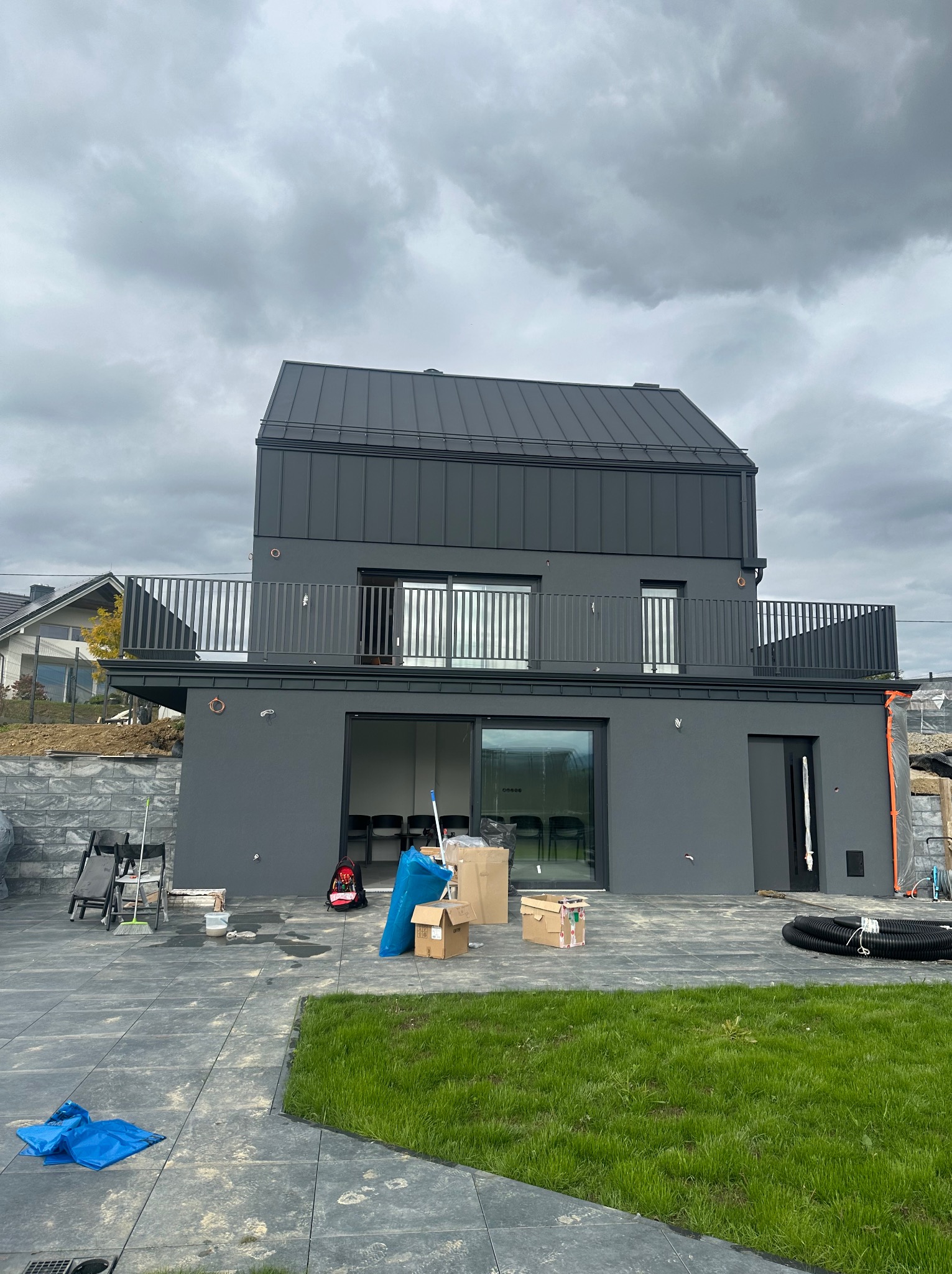 Dwupiętrowy dom z szarą elewacją i metalowym dachem w pochmurny dzień. Widoczne elementy wykończeniowe i narzędzia na zewnątrz budynku.