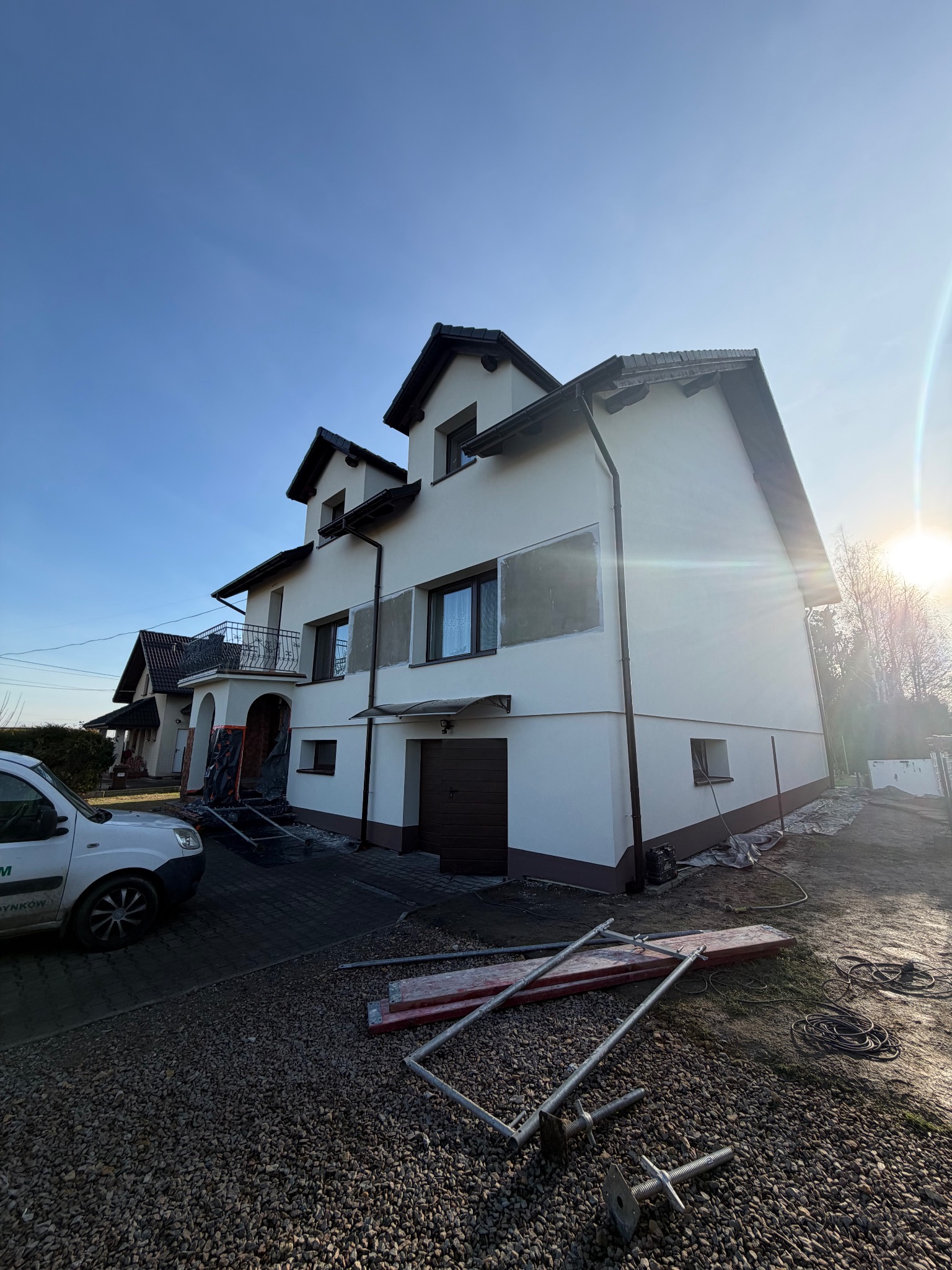 Dom jednorodzinny z fragmentami elewacji przygotowanymi do malowania, widoczne rusztowania i samochód dostawczy firmy budowlanej. Słoneczny dzień.