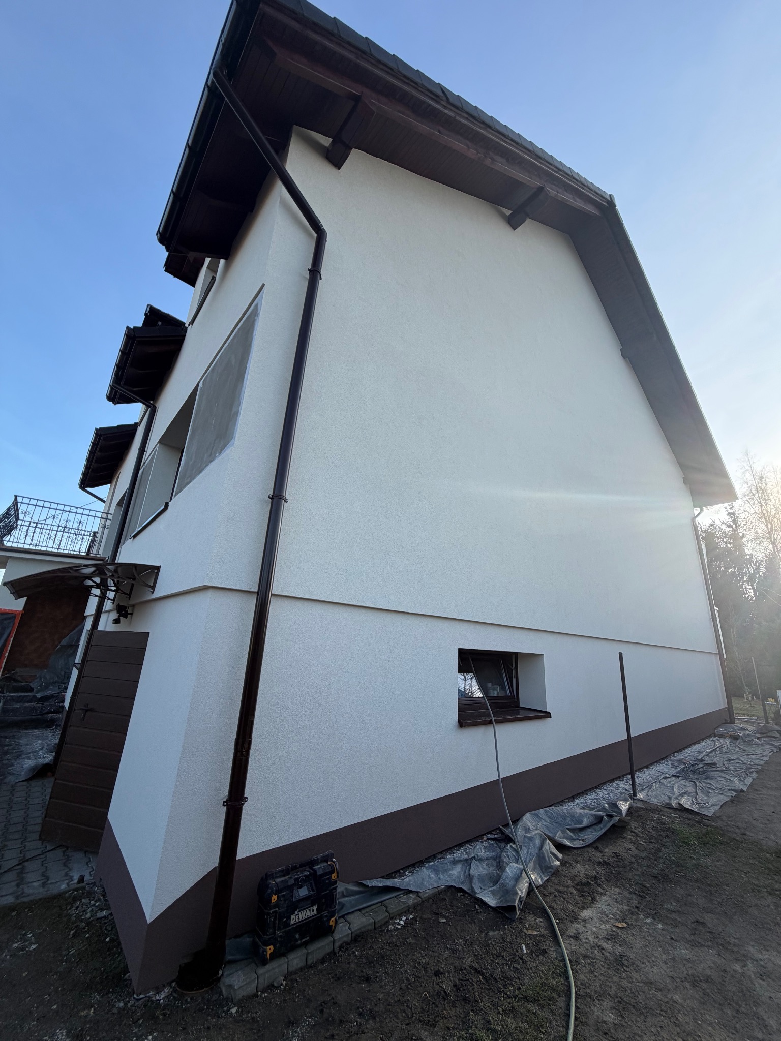 Boczny widok domu po tynkowaniu elewacji na biało z brązowymi akcentami. Widoczne okna, rynna, narzędzia i folia ochronna na ziemi. Słoneczny dzień.