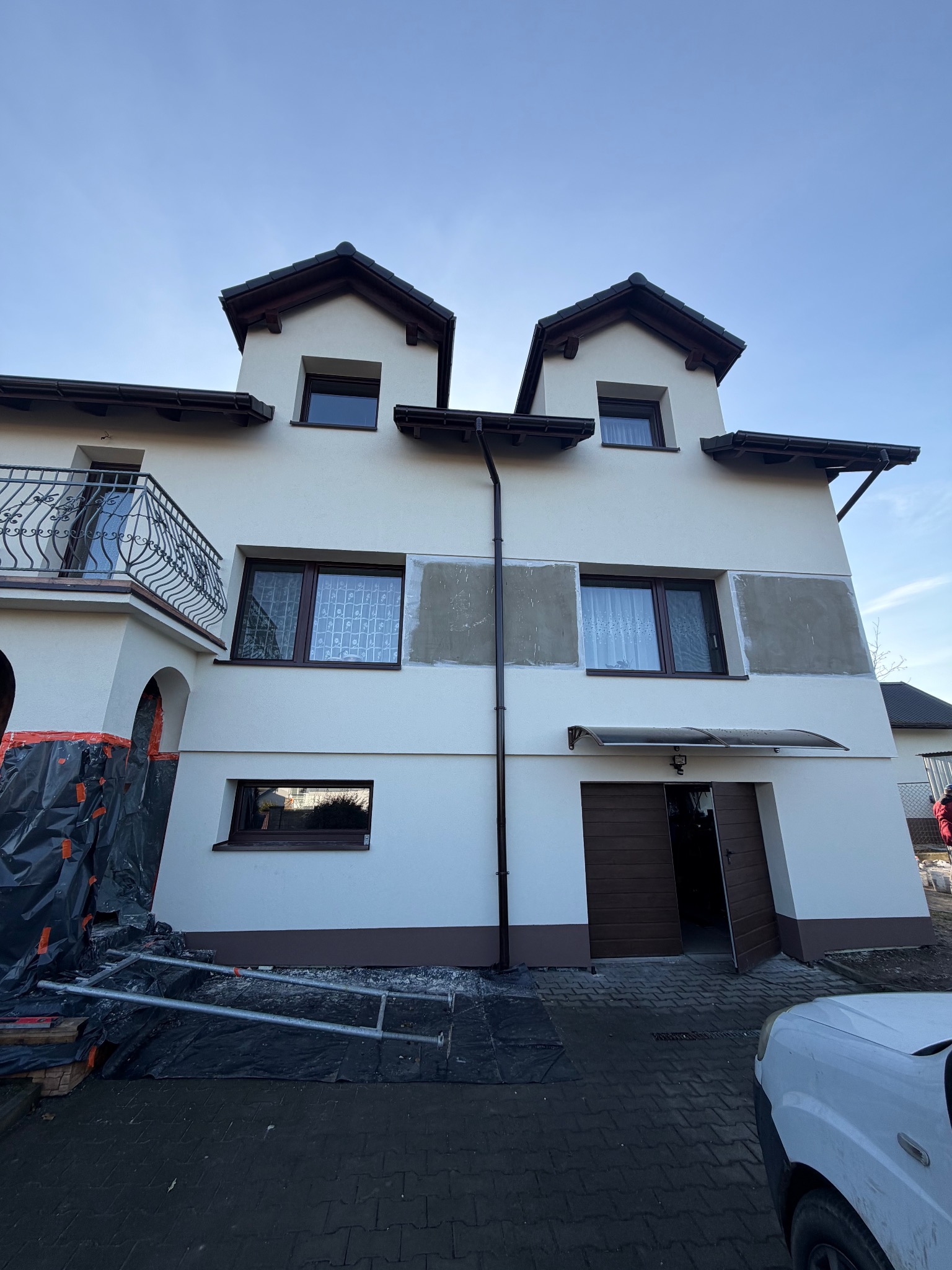 Dwupiętrowy dom z białymi ścianami i brązowymi akcentami, w trakcie prac elewacyjnych. Widoczne rusztowania i zabezpieczenia. Fragmenty ścian przygotowane do malowania.