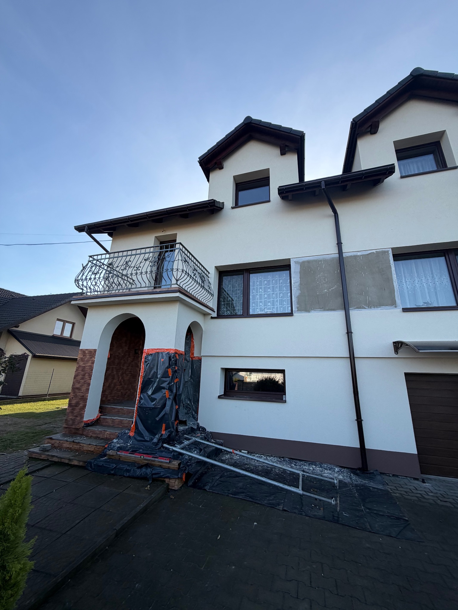 Dwupiętrowy dom z balkonem w trakcie prac elewacyjnych. Fragment ściany bez tynku, zabezpieczone wejście, rusztowanie. Słoneczny dzień.