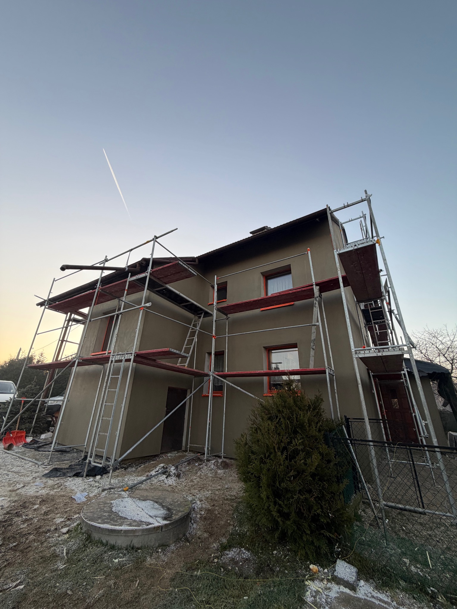 Dom jednorodzinny w trakcie tynkowania elewacji, otoczony rusztowaniami. Widoczny fragment nieba z smugą kondensacyjną samolotu. Ujęcie z dołu.