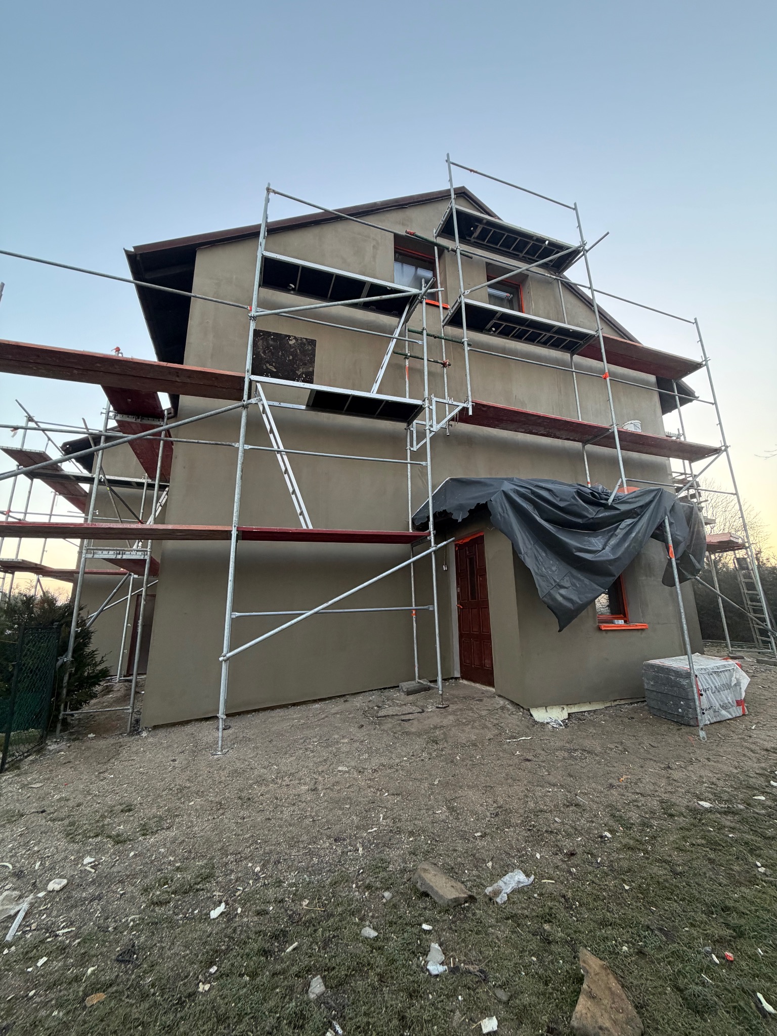 Dwupiętrowy dom w trakcie tynkowania elewacji, otoczony rusztowaniami. Widoczne okna z pomarańczowymi ramami i czarna plandeka nad drzwiami. Na ziemi leżą materiały budowlane.
